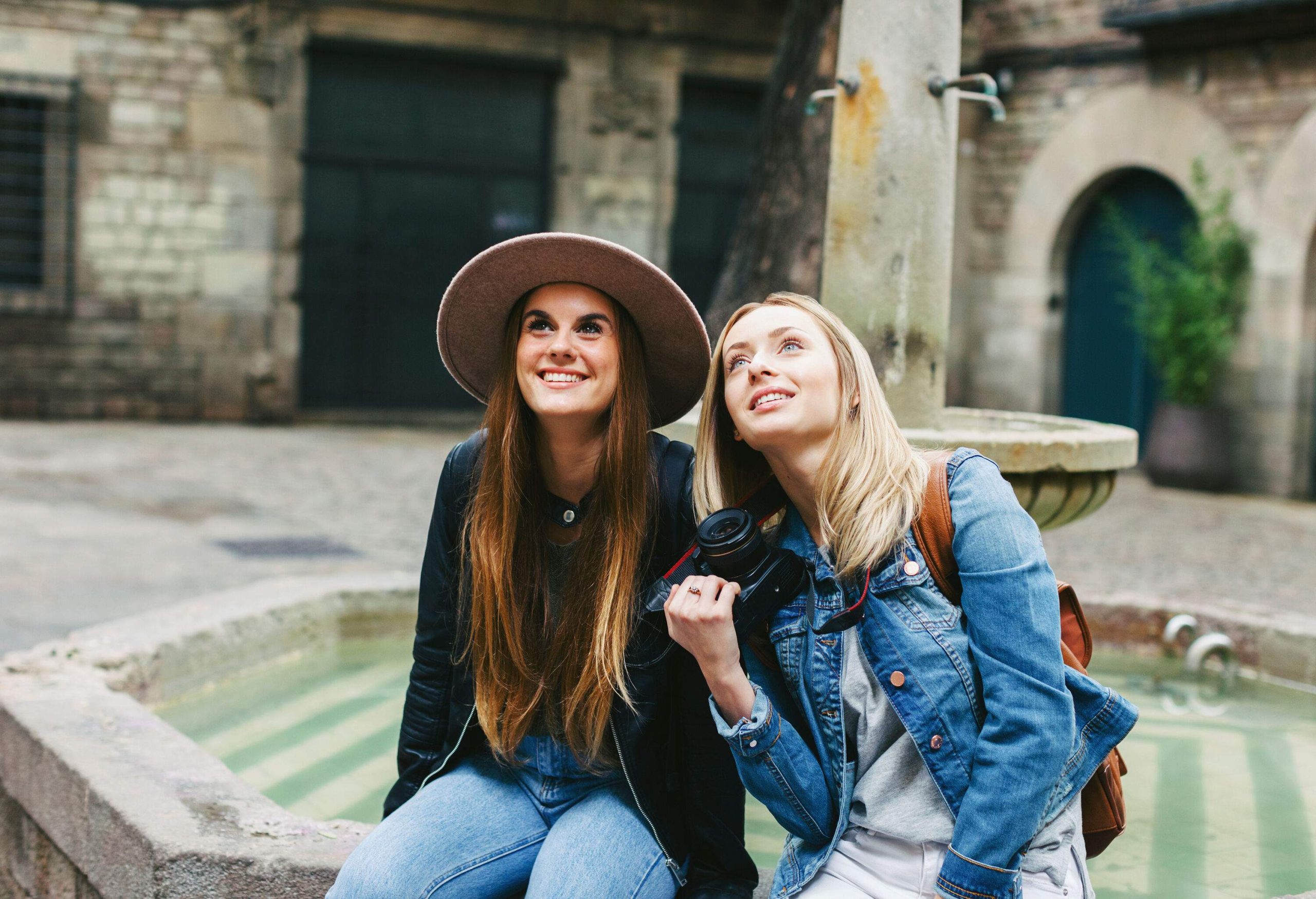 Two young women sit at a fountain, gazing upwards, one wearing a gorgeous hat, and both dressed comfortably in jackets.