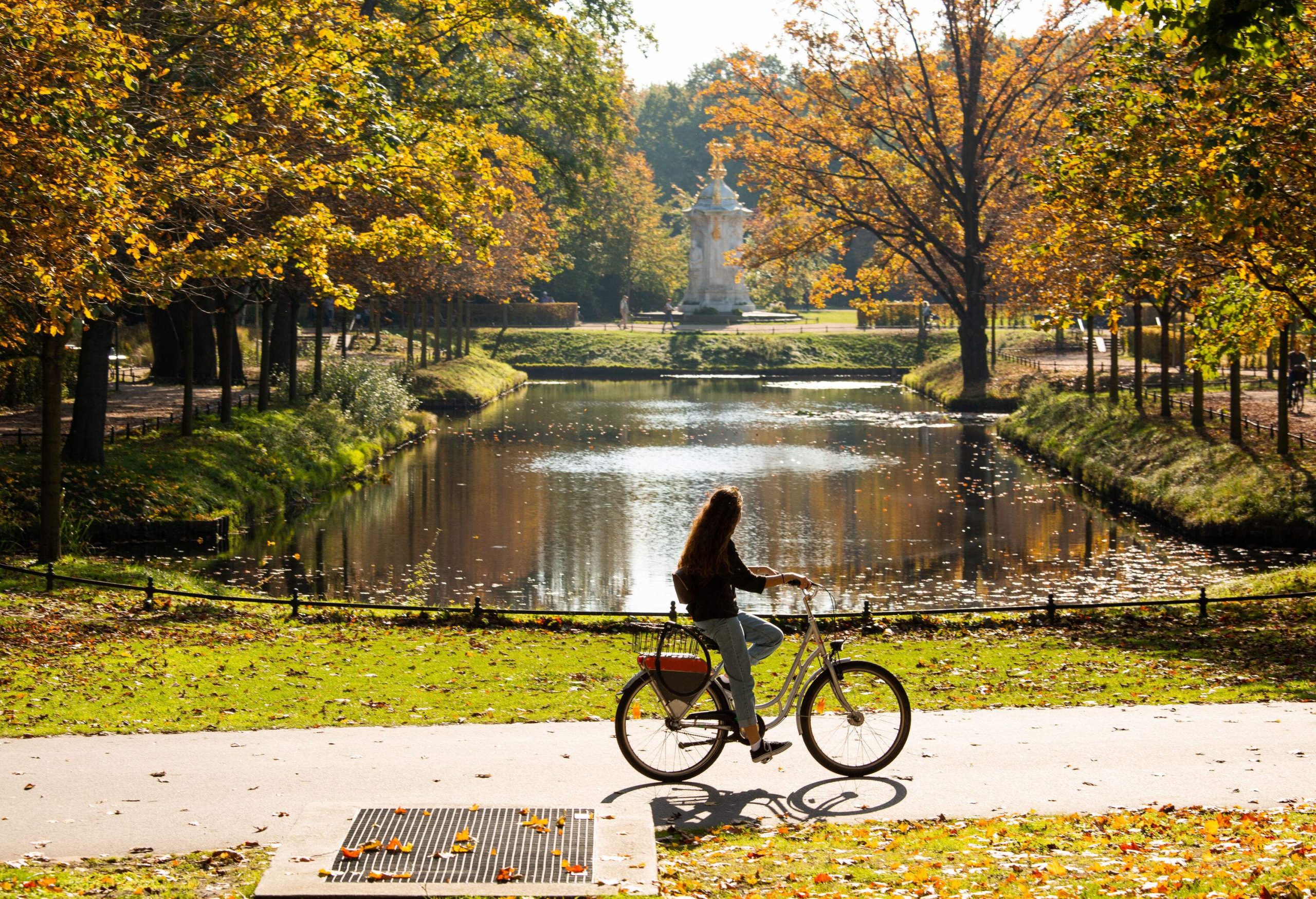 A small pond surrounded by lush foliage and a woman cycling along the park's paved trails.