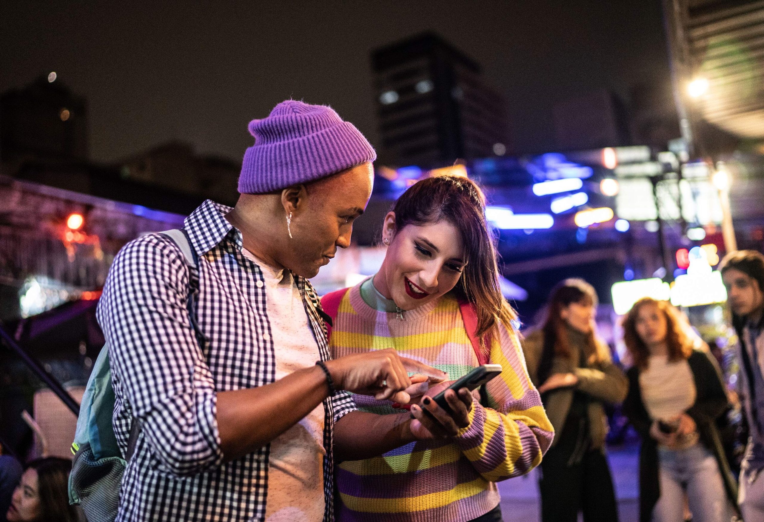 A couple looking over at something on a smartphone while they stand on a street.