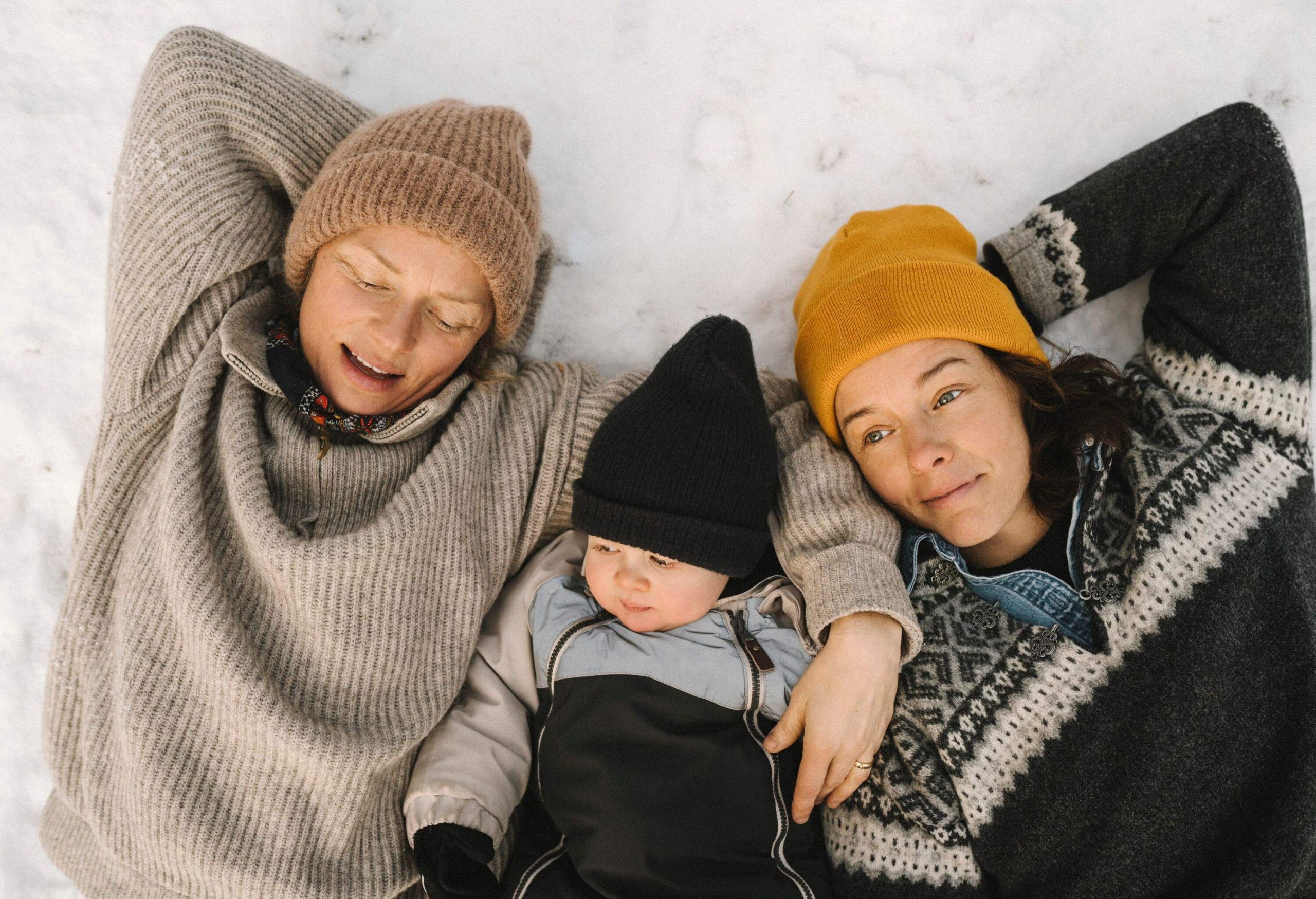 A lesbian LGBTQ couple and their daughter, wearing hats, are lying in the snow