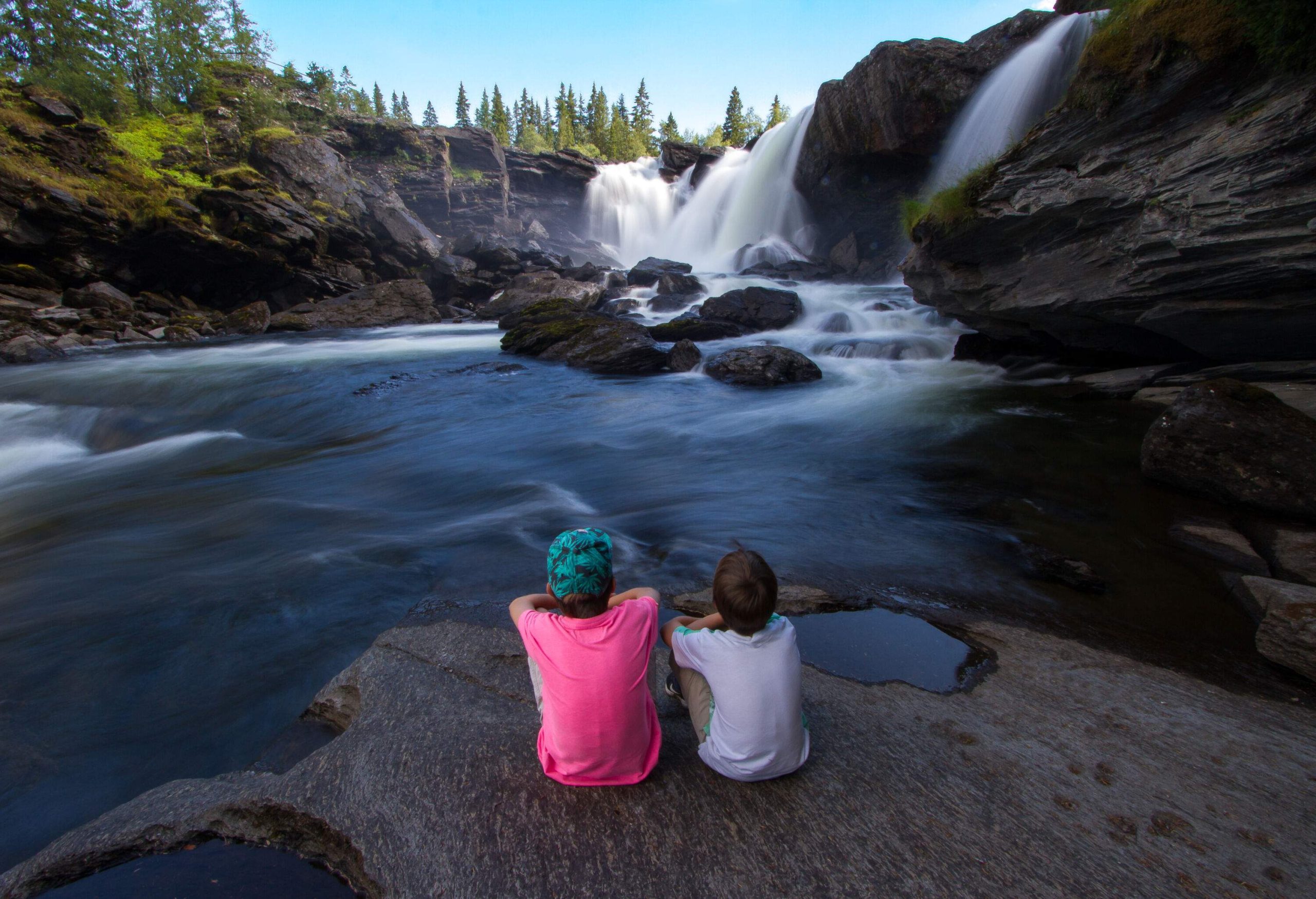 Two kids sit on a flat rock admiring a waterfall that flows into a rocky river.