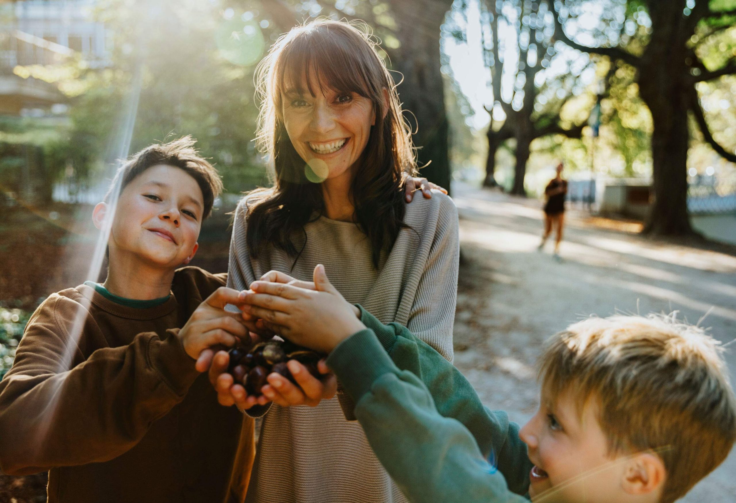 A mature woman and two young boys collecting chestnuts on a crisp autumn day.