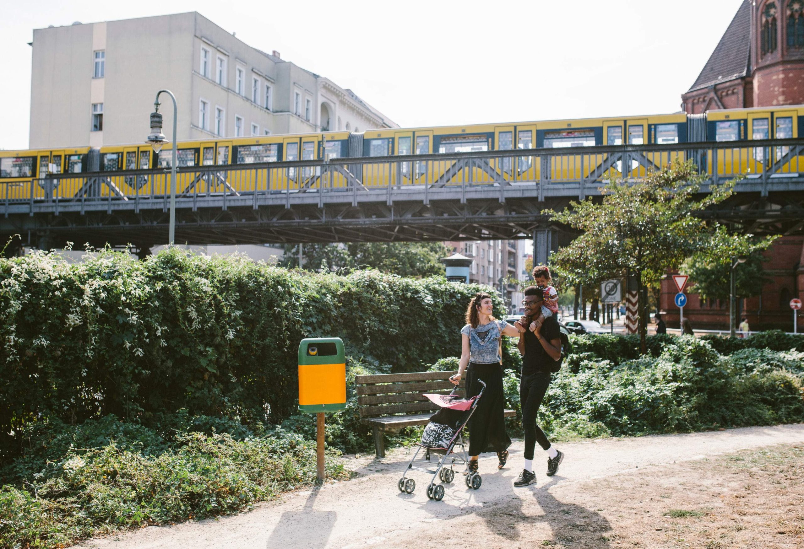 A father carries his toddler on his shoulder alongside her wife, walking on an unpaved pathway with a yellow train travelling above them in the background.