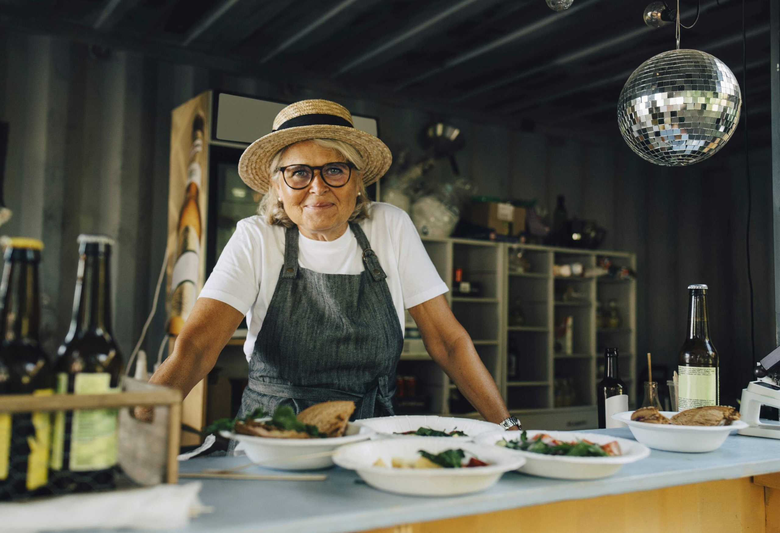 An elderly woman in a straw hat and apron stands beside a restaurant counter full of food.