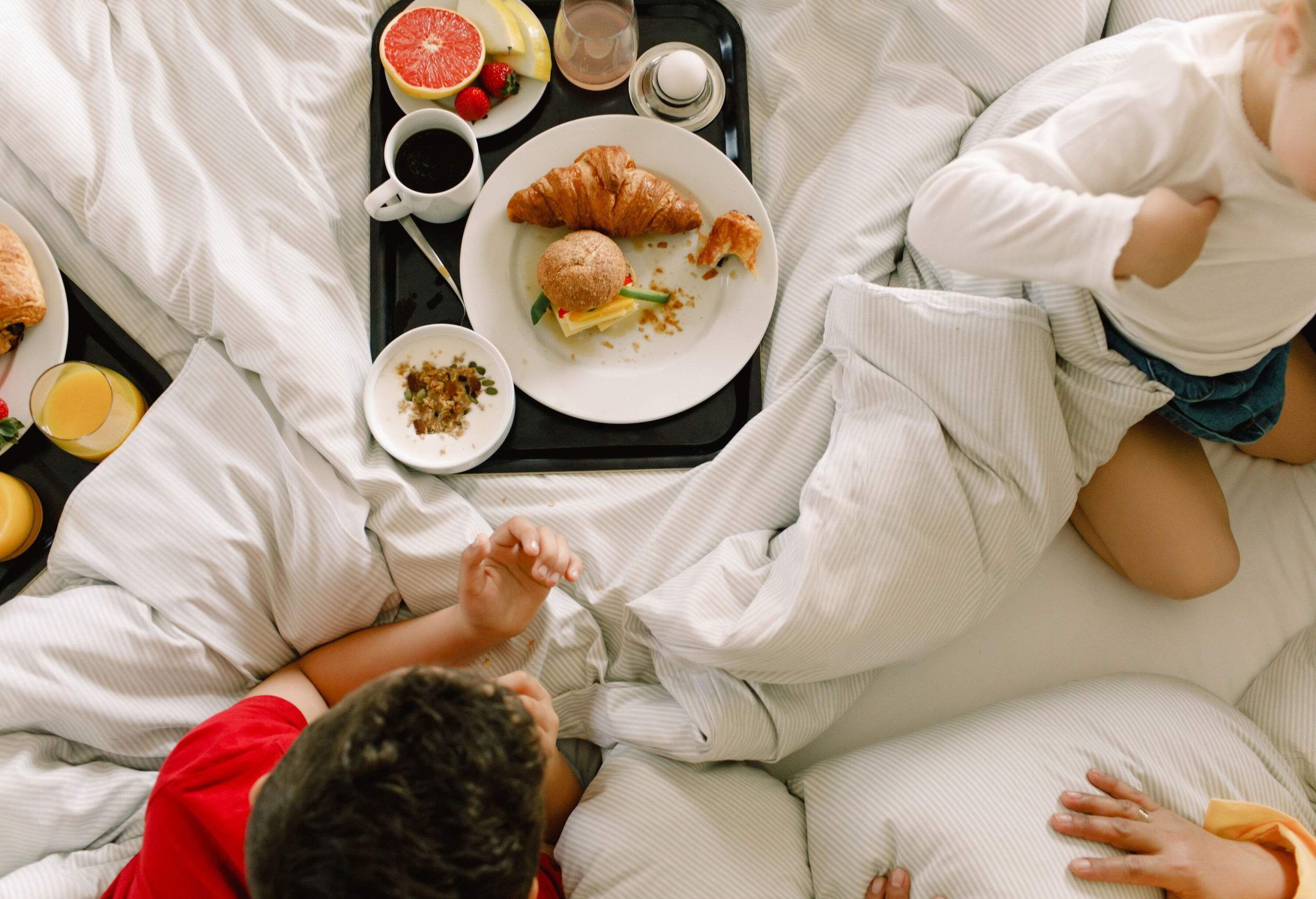 A group of kids on the bed with a tray of snacks including a plate of pastries and fresh fruits, a cup of coffee, and a bowl of cereal.