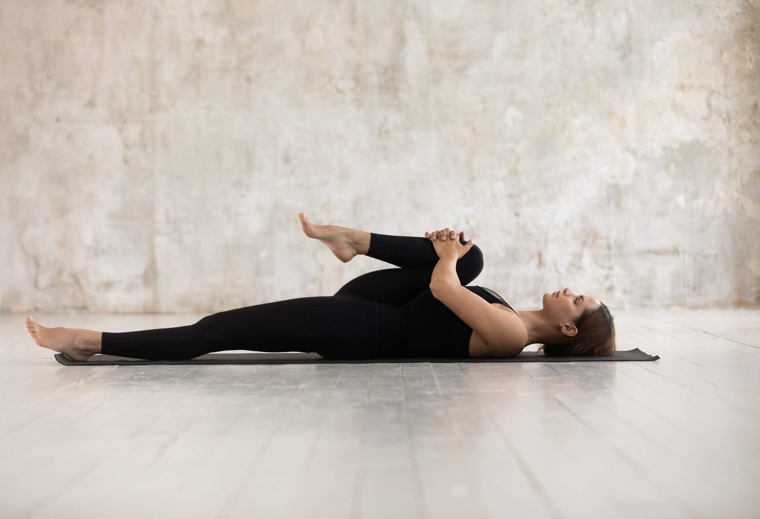 A woman dressed in all-black exercise attire lying on the floor with one knee pressed against her chest.