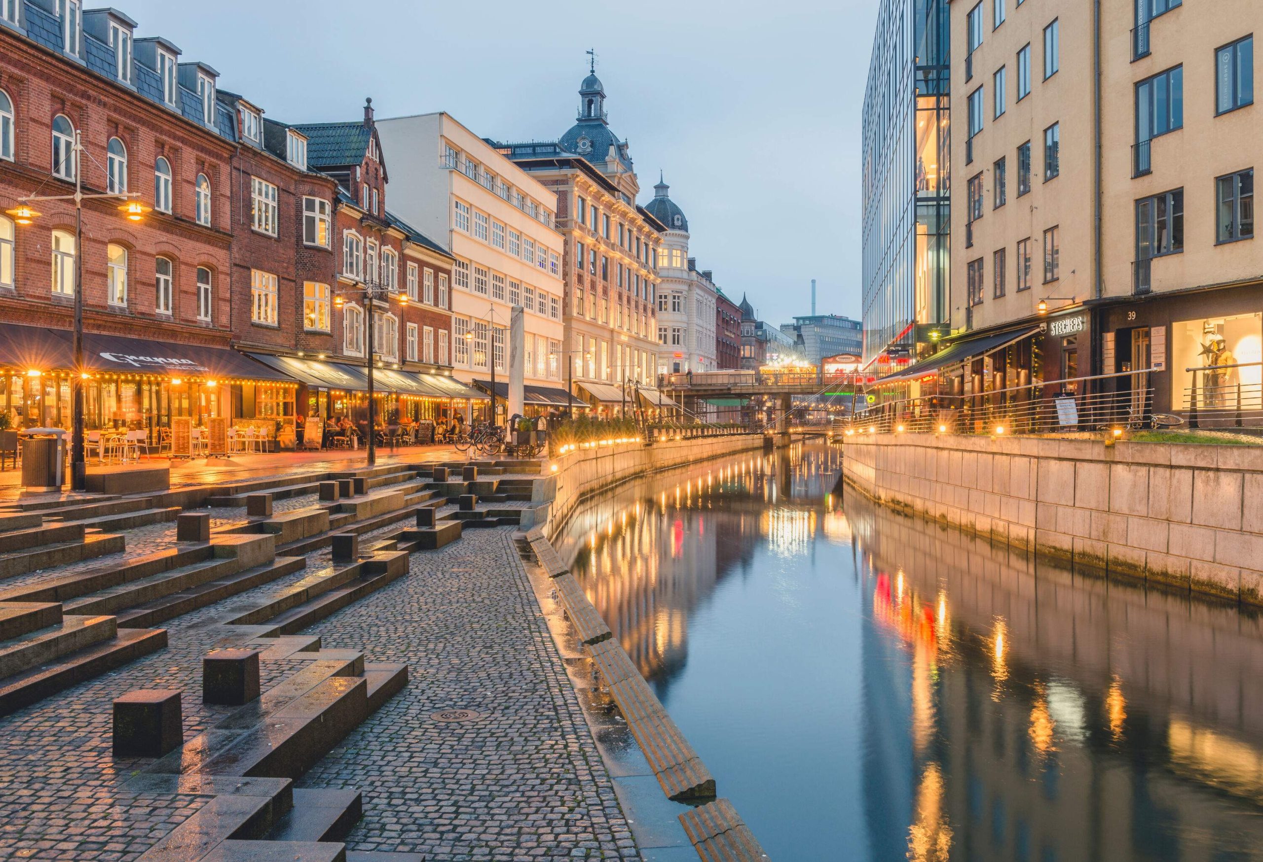 A night-time view of a river canal along a promenade that reflects the surrounding buildings with bright lights.