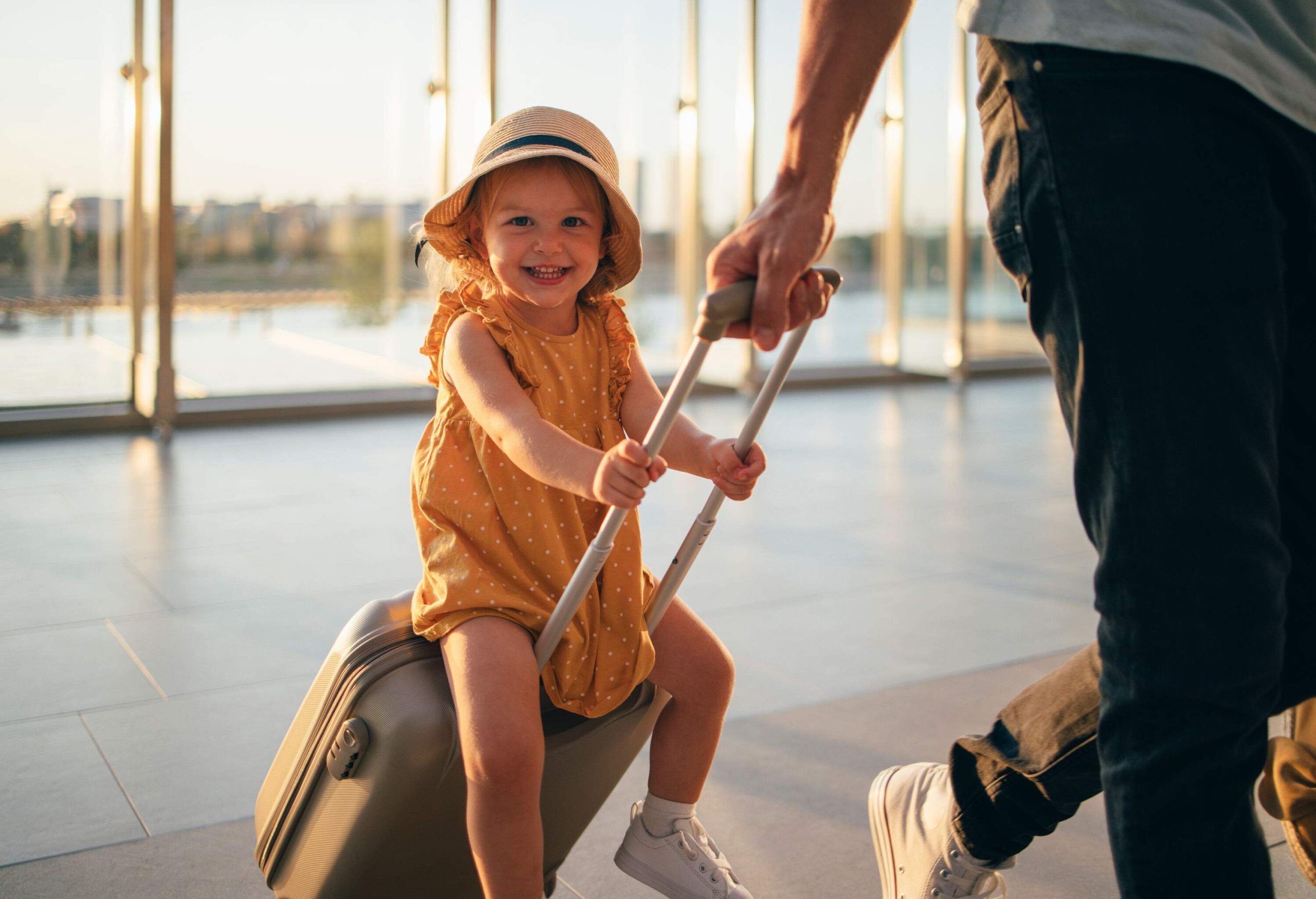 A little girl having fun riding atop a suitcase as a man drags it across the tiled floor.