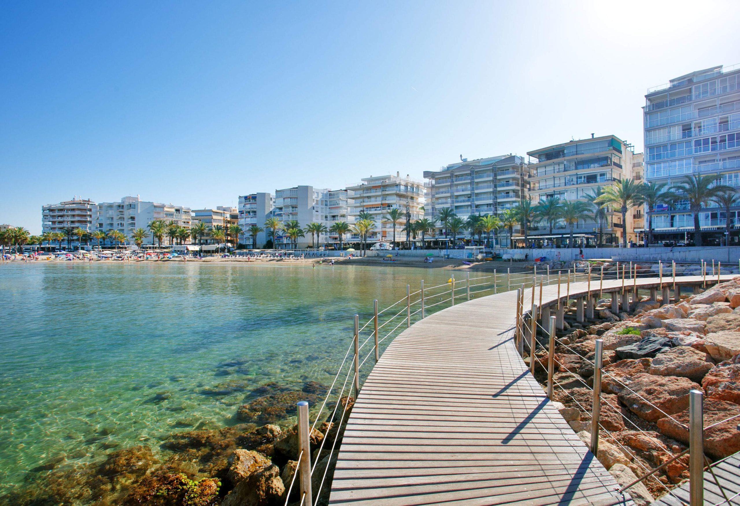 A boardwalk runs across the crystal-clear water to the coastline, where it connects to a complex of multi-storey buildings and a palm tree-lined coastal road.