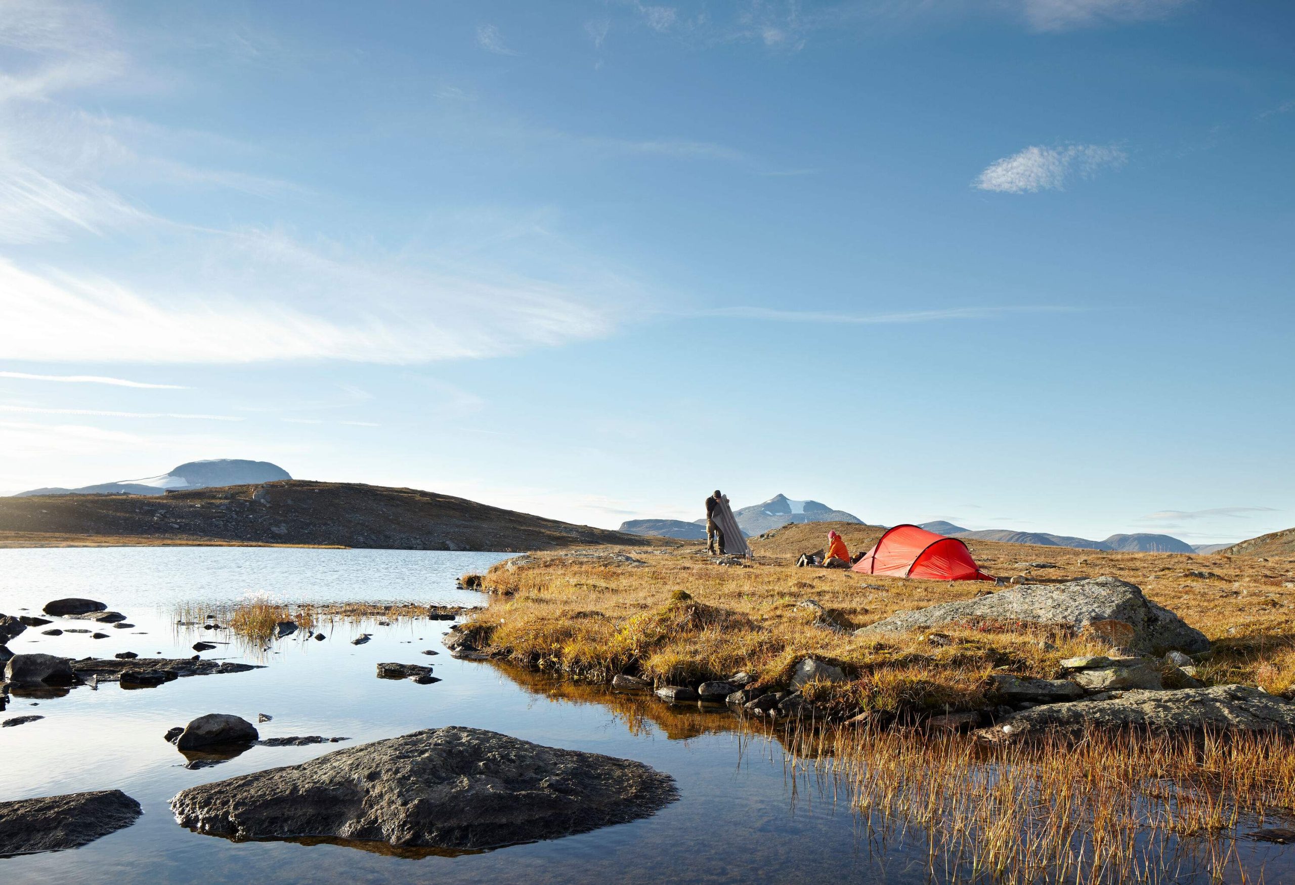 A man and woman wild-camping near a mountain lake with a red tunnel tent.