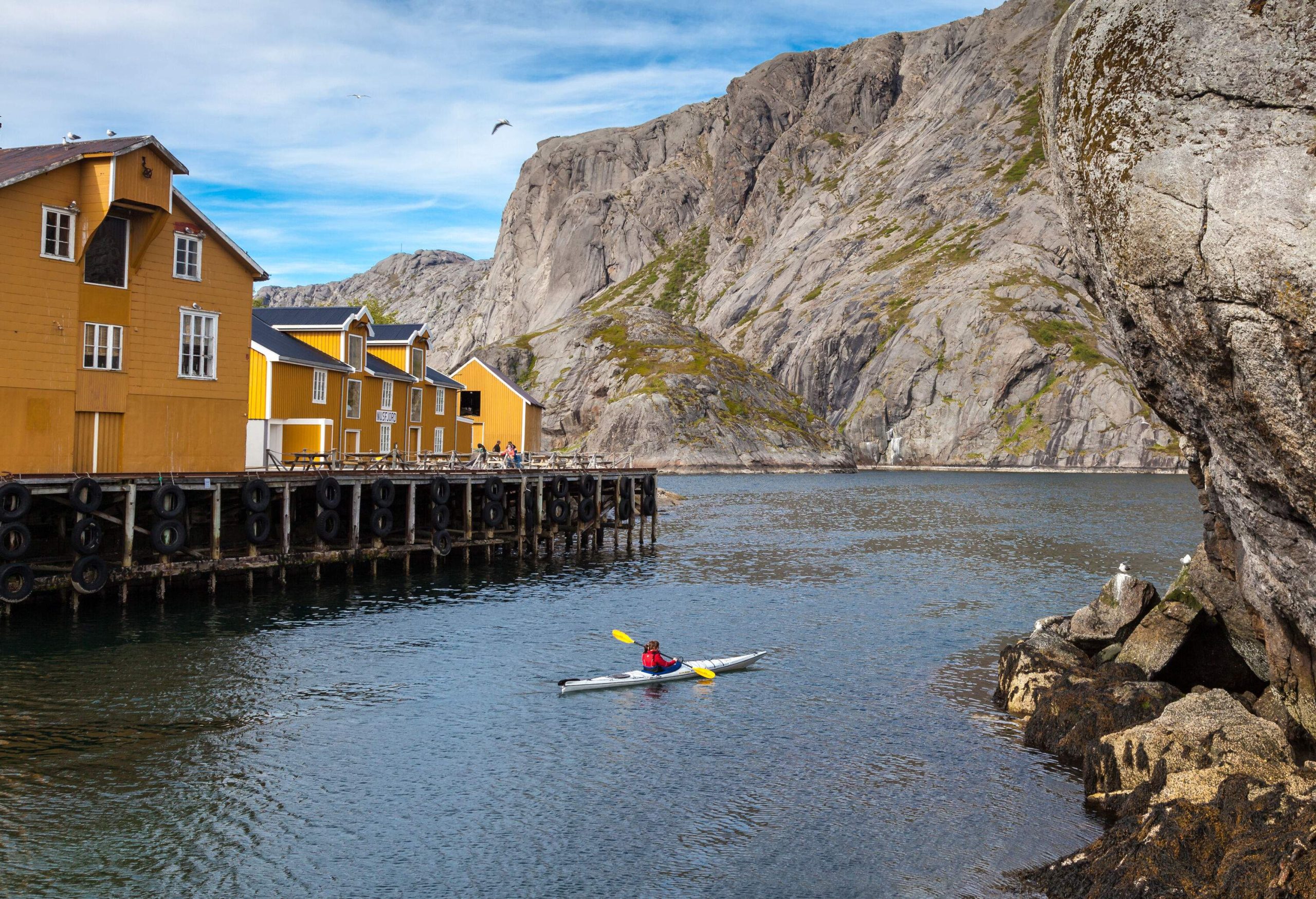 A man paddling a kayak in a fjord along a tranquil yellow fishing village bounded by steep rock mountains.