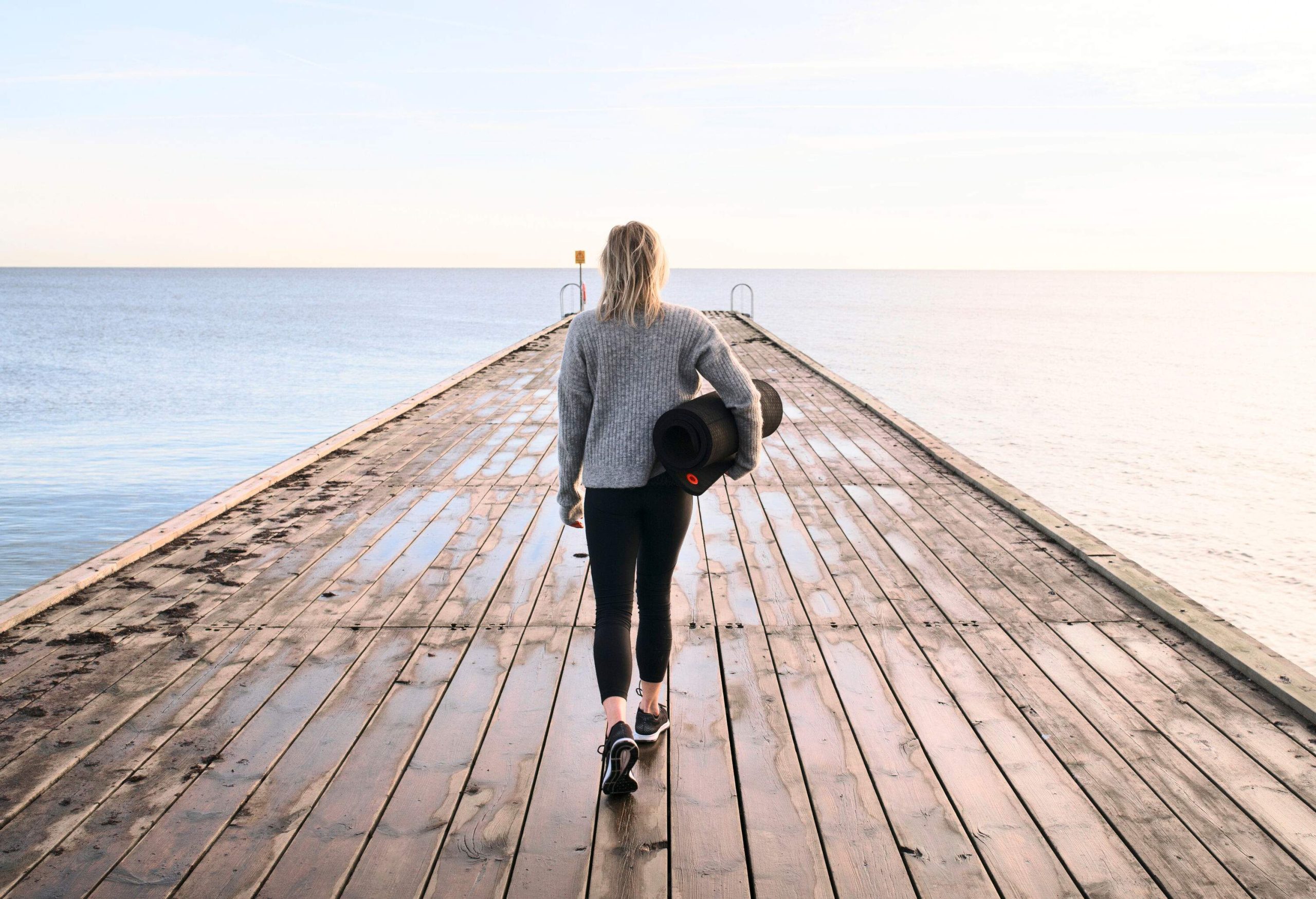 A woman in grey sweater carries a mat as she walks across a wooden pier fronting a vast ocean.