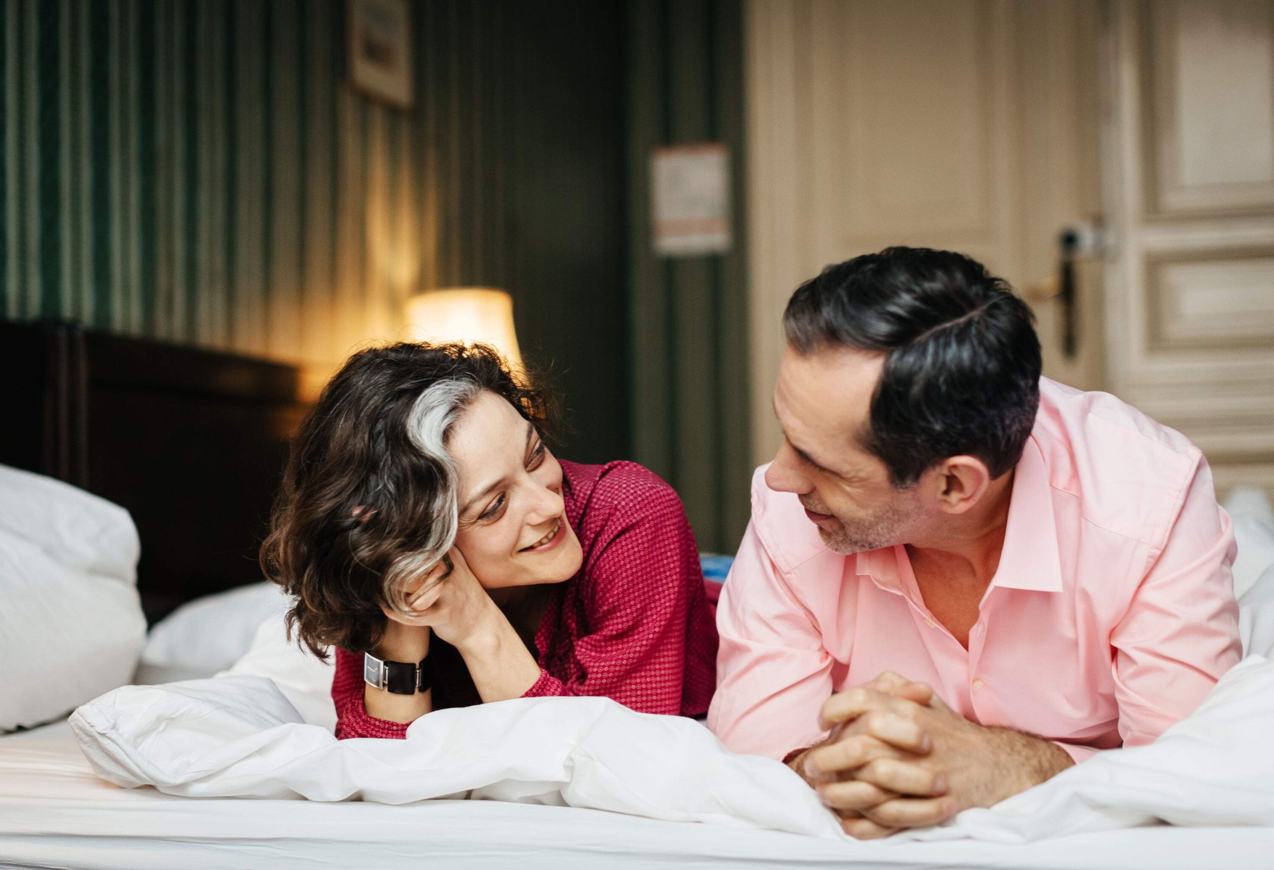 A mature, happy couple is lying at a hotel bed next to each other. They both are smiling and seem to be in love. In the background, the dark and green interior of the room can be seen.