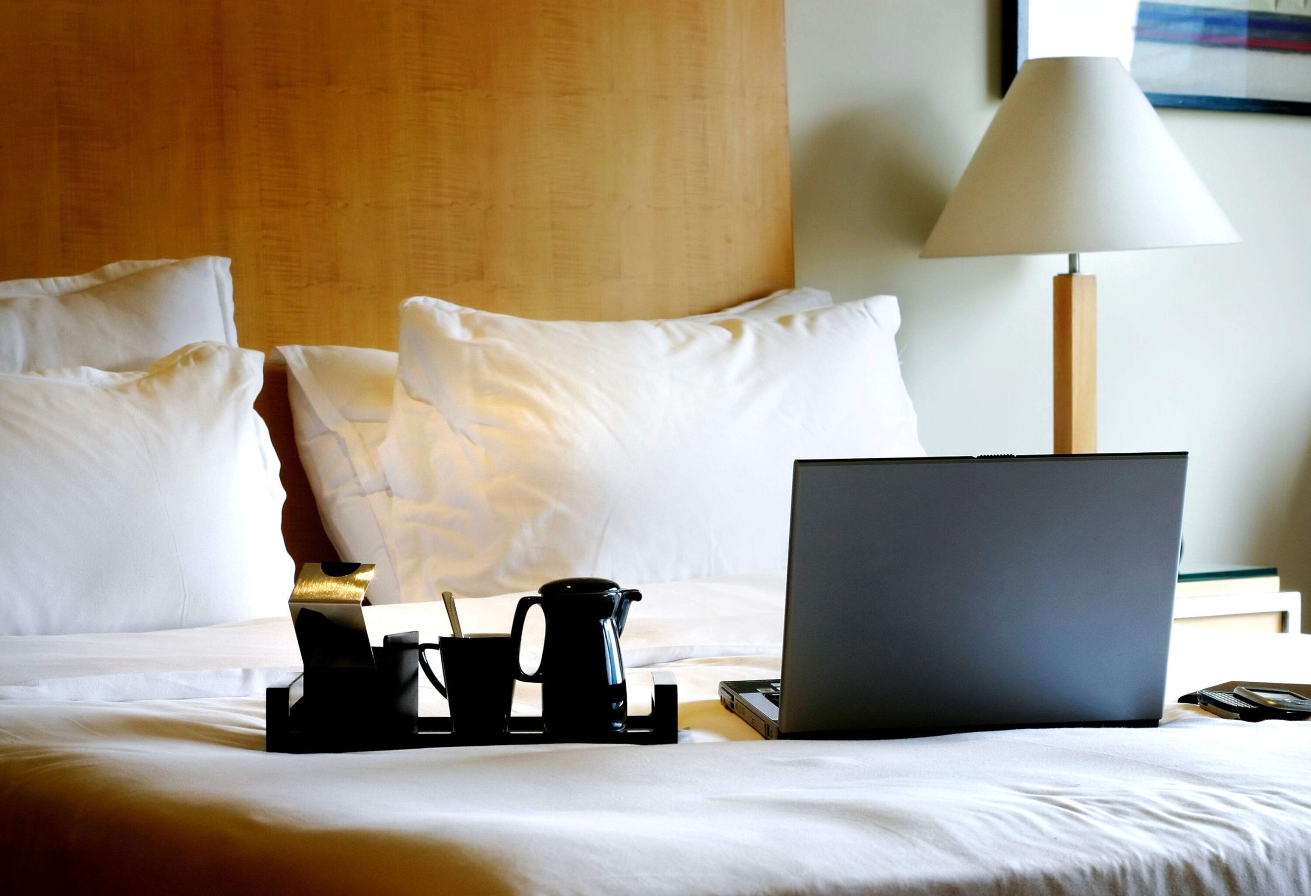A laptop and black coffee ware in a white bed with a wooden headboard.