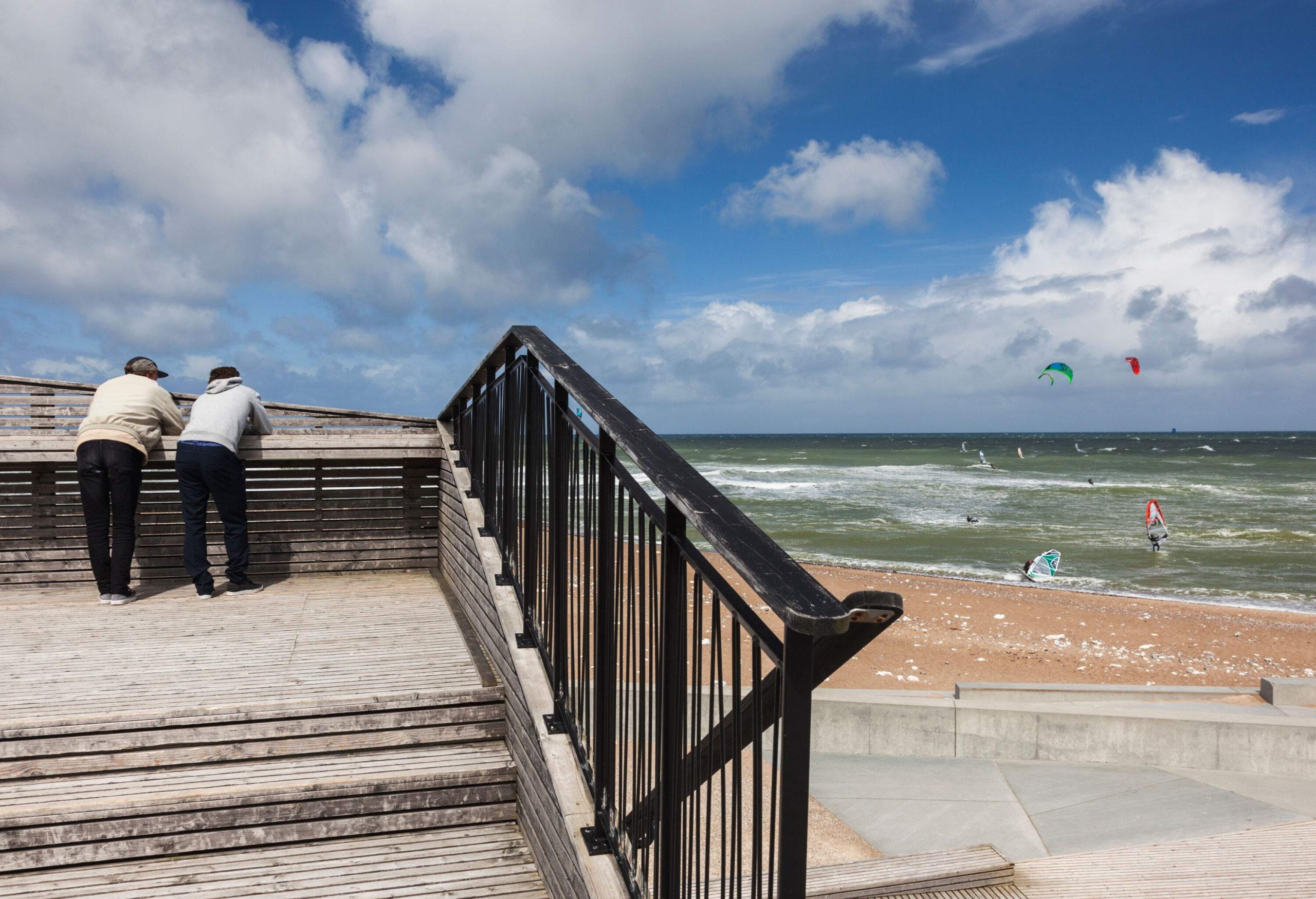 Two people leaning on a terrace's wooden railing, watching people windsurf in the ocean.