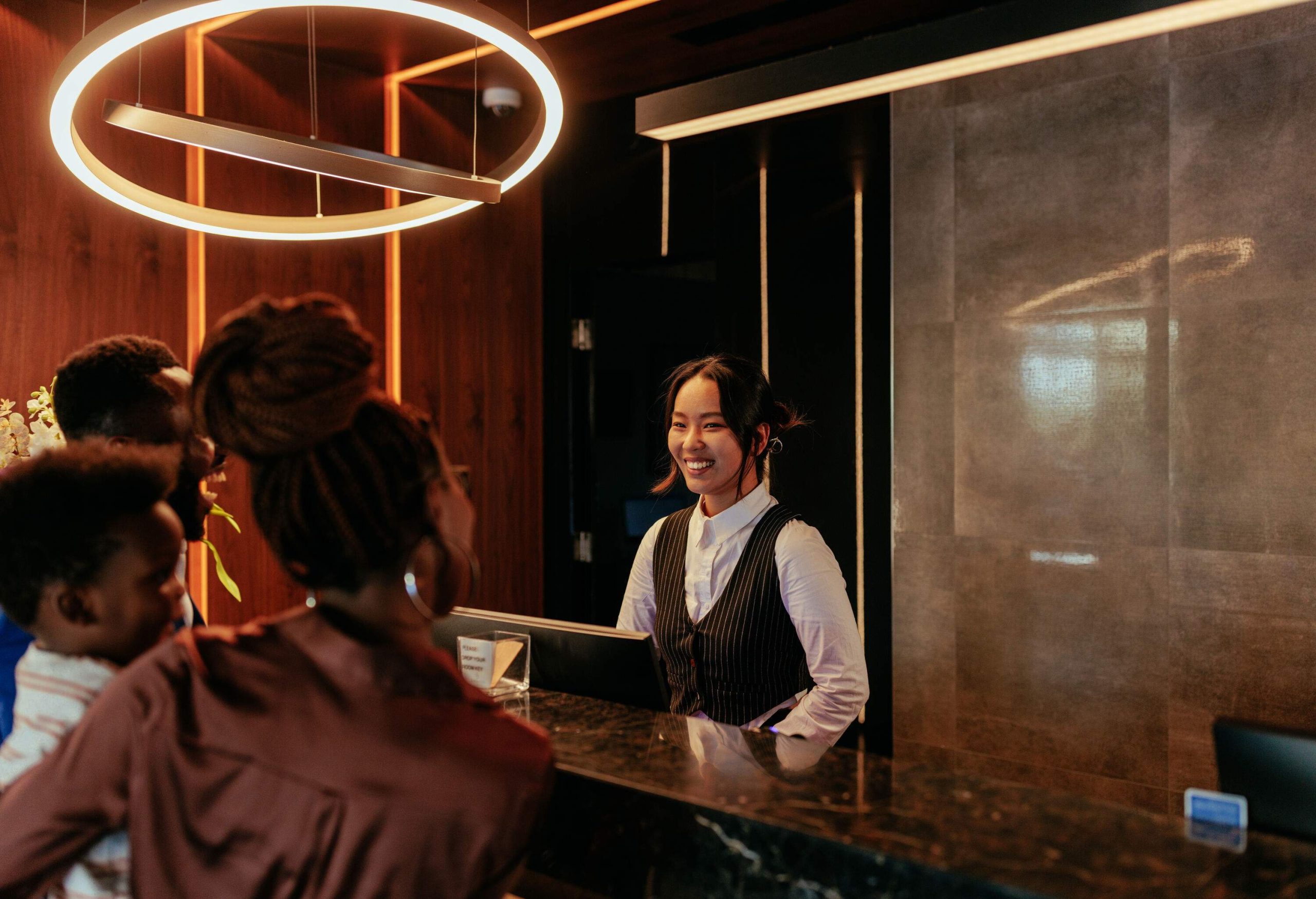A young receptionist is at the front desk of a luxurious hotel talking to her African American hotel guests.