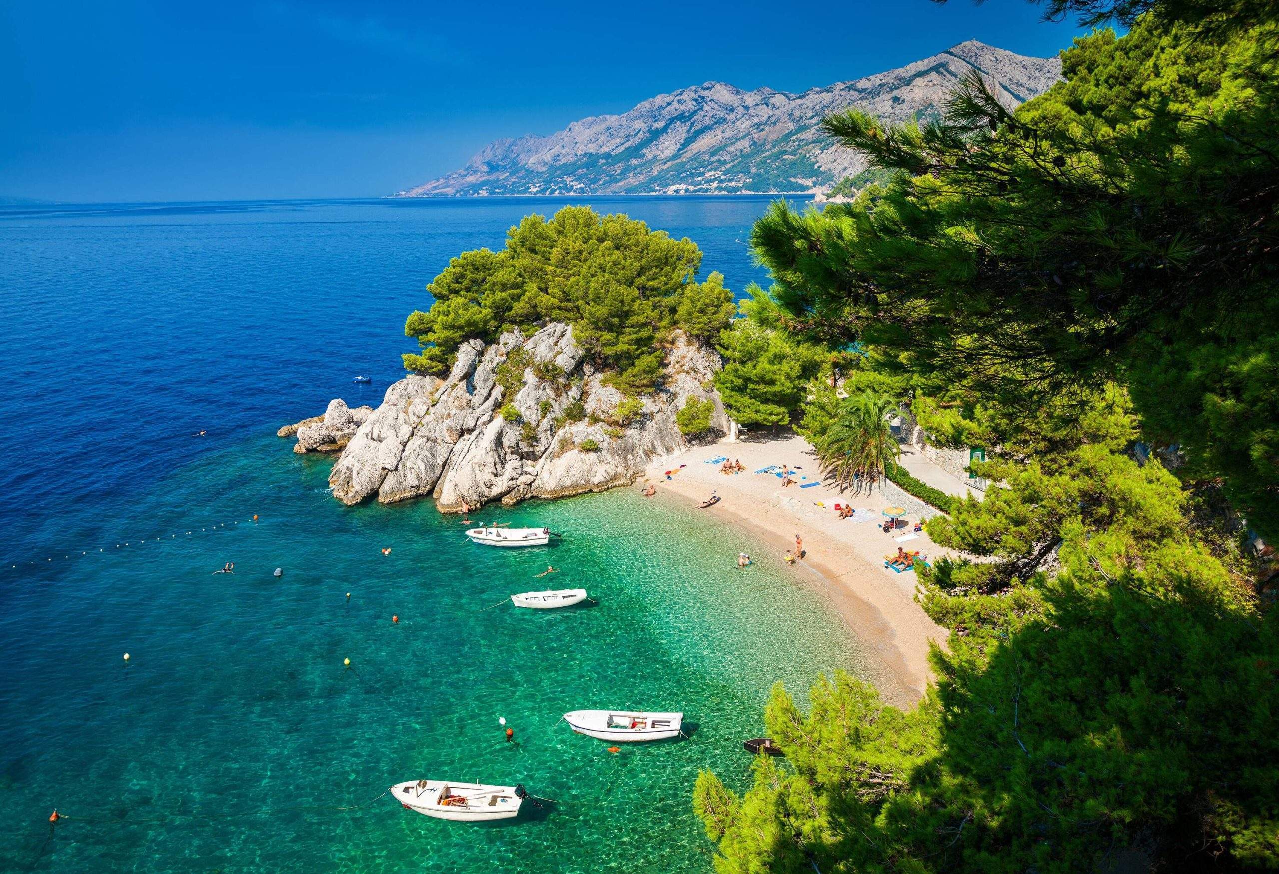 Boats anchored on a turquoise beach with massive rocks covered in vegetation.