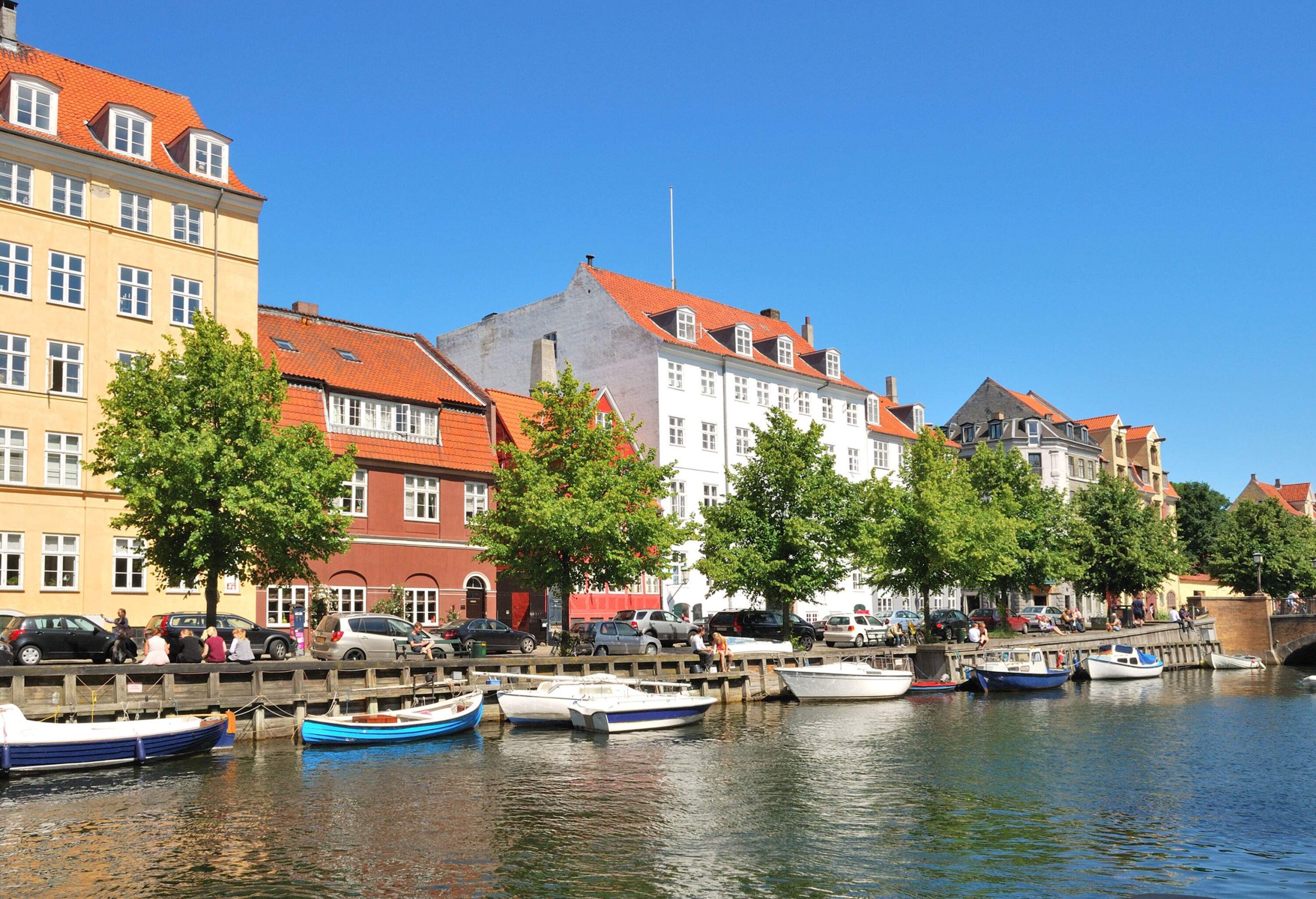 Several small boats lined up on a canal alongside a road with parked cars in front of colourful buildings.