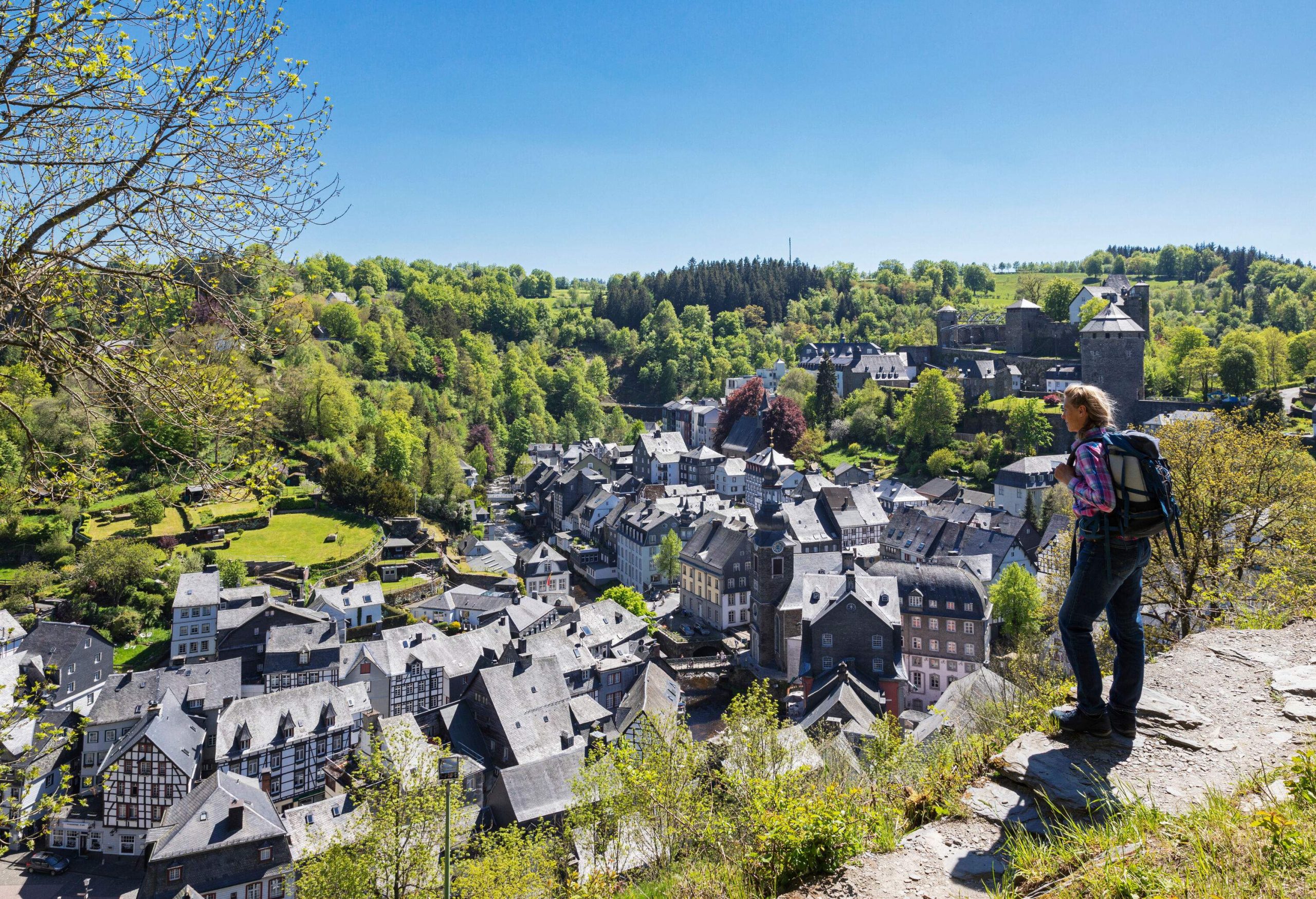 A hiker on a cliffside path overlooking a village surrounded by dense forest.