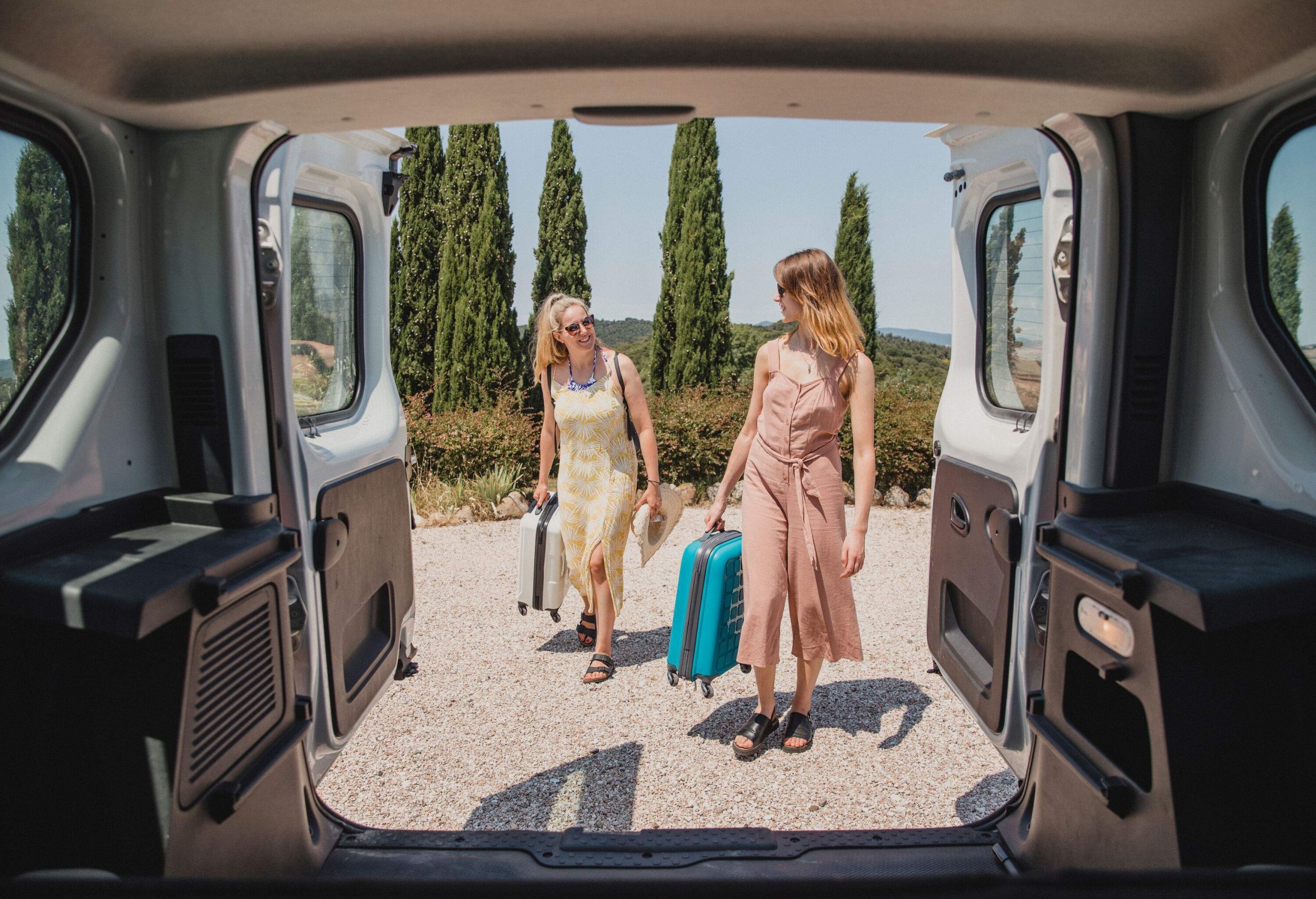 Two women look at each other while loading their luggage on a car's trunk.