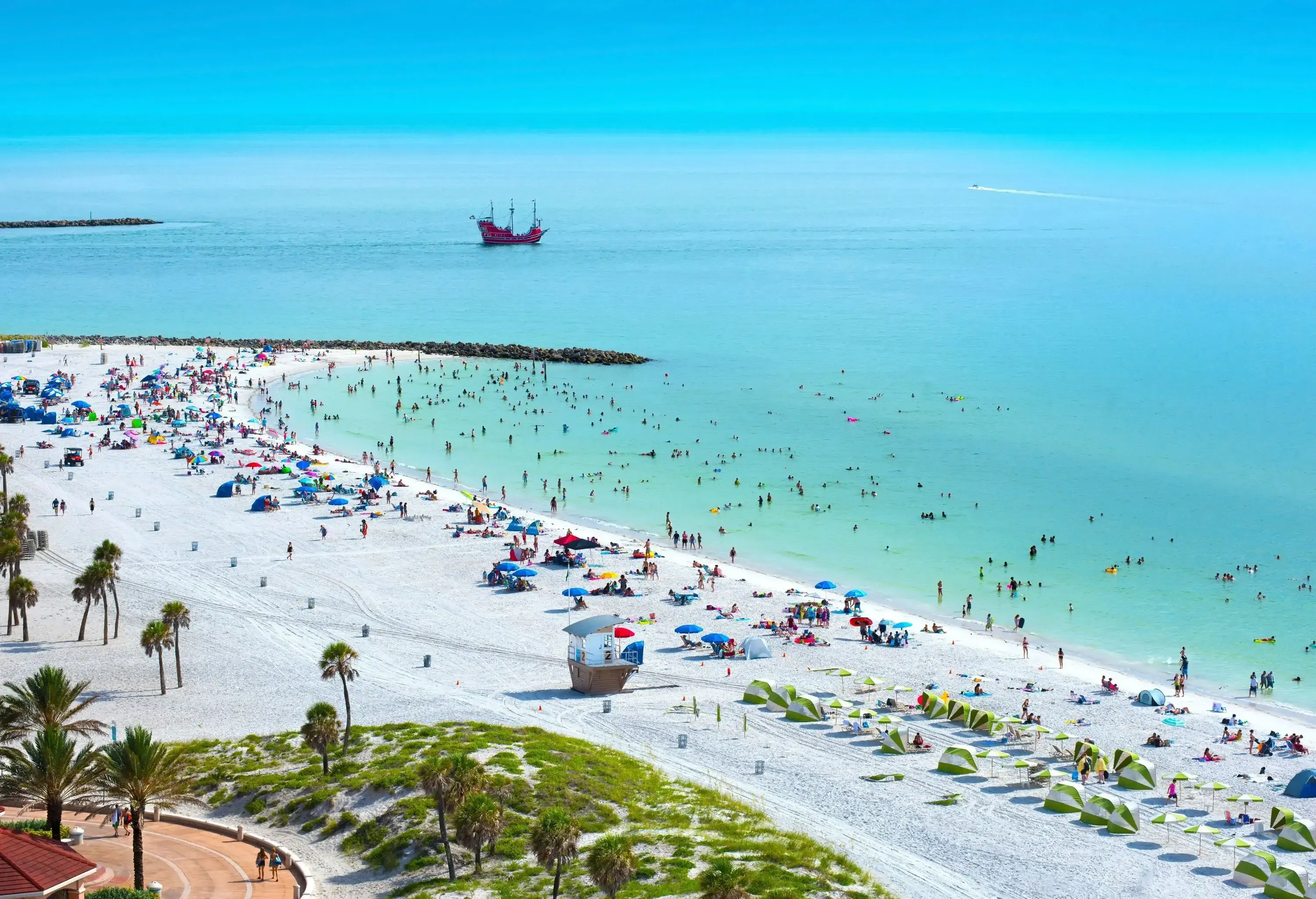 A bustling Clearwater Beach teems with visitors as colourful parasols and sunbeds dot the shoreline, while the sea shimmers in the sun and a sailing ship sails majestically in the distance.