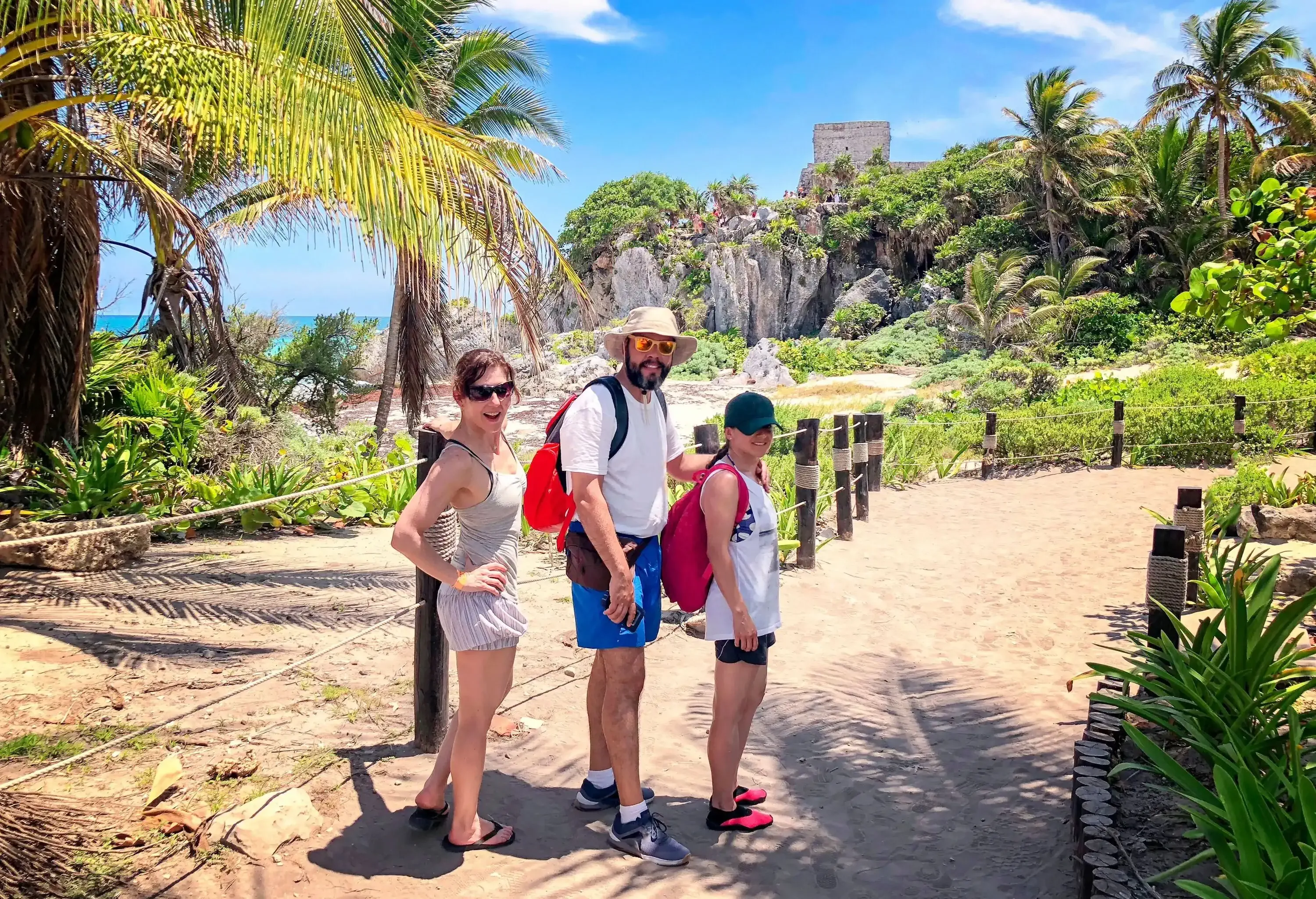 A radiant family of three, donning joyful smiles, walks along a sandy path, framed by lush tropical trees that provide a serene backdrop.