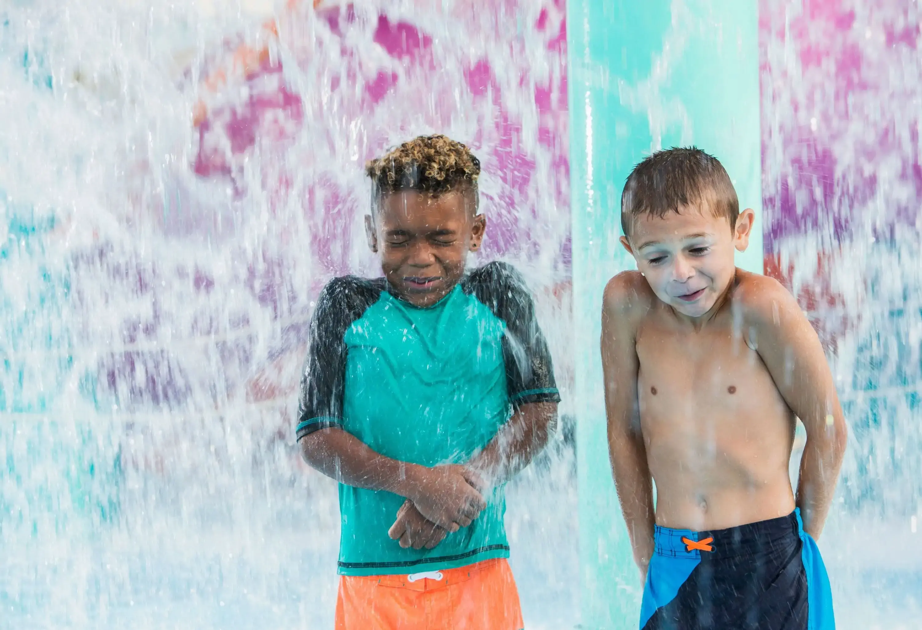 Two young boys getting soaked while standing under a waterpark waterfall.