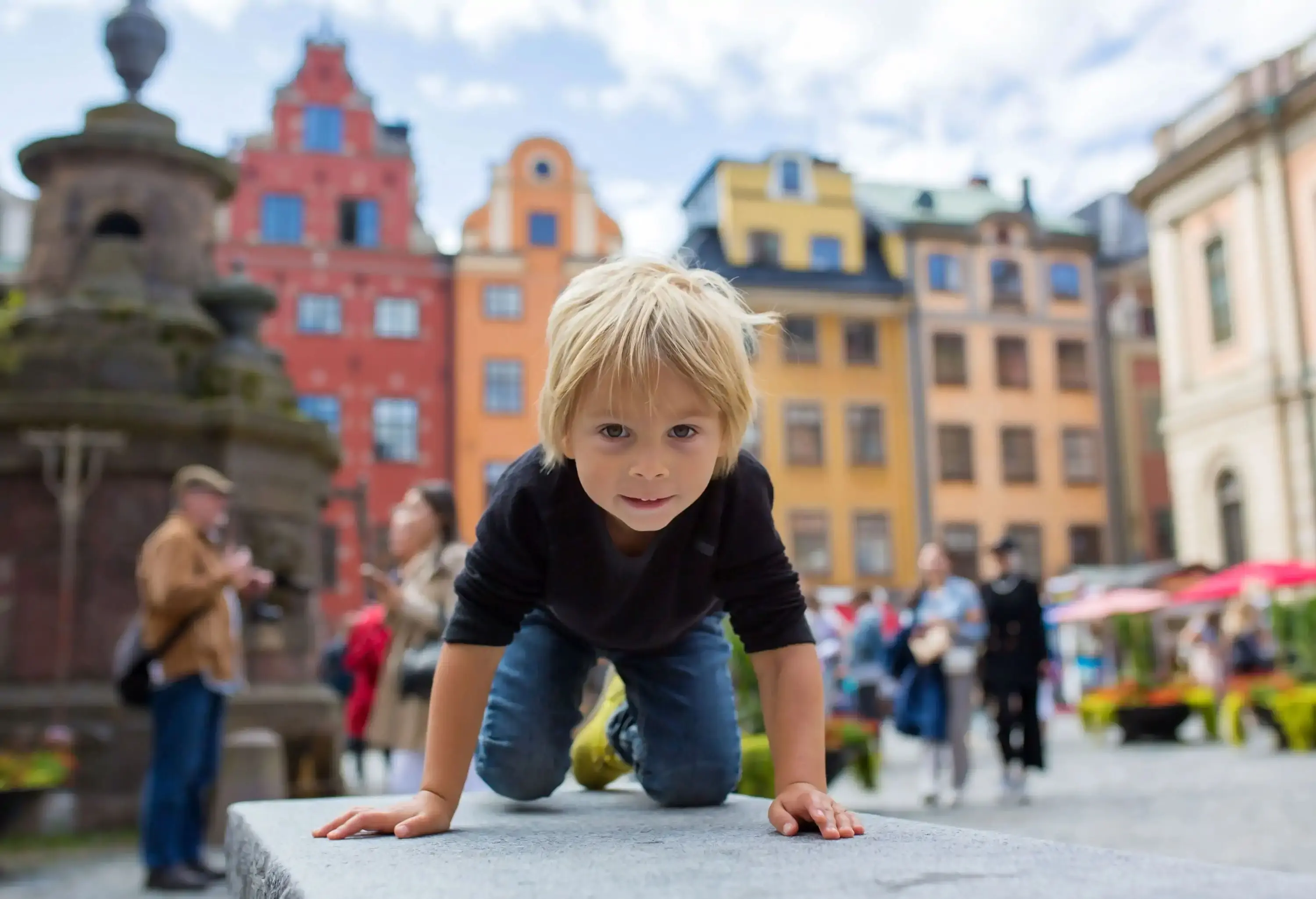 A kid crawling on a bench in a plaza with colourful buildings and a water fountain.