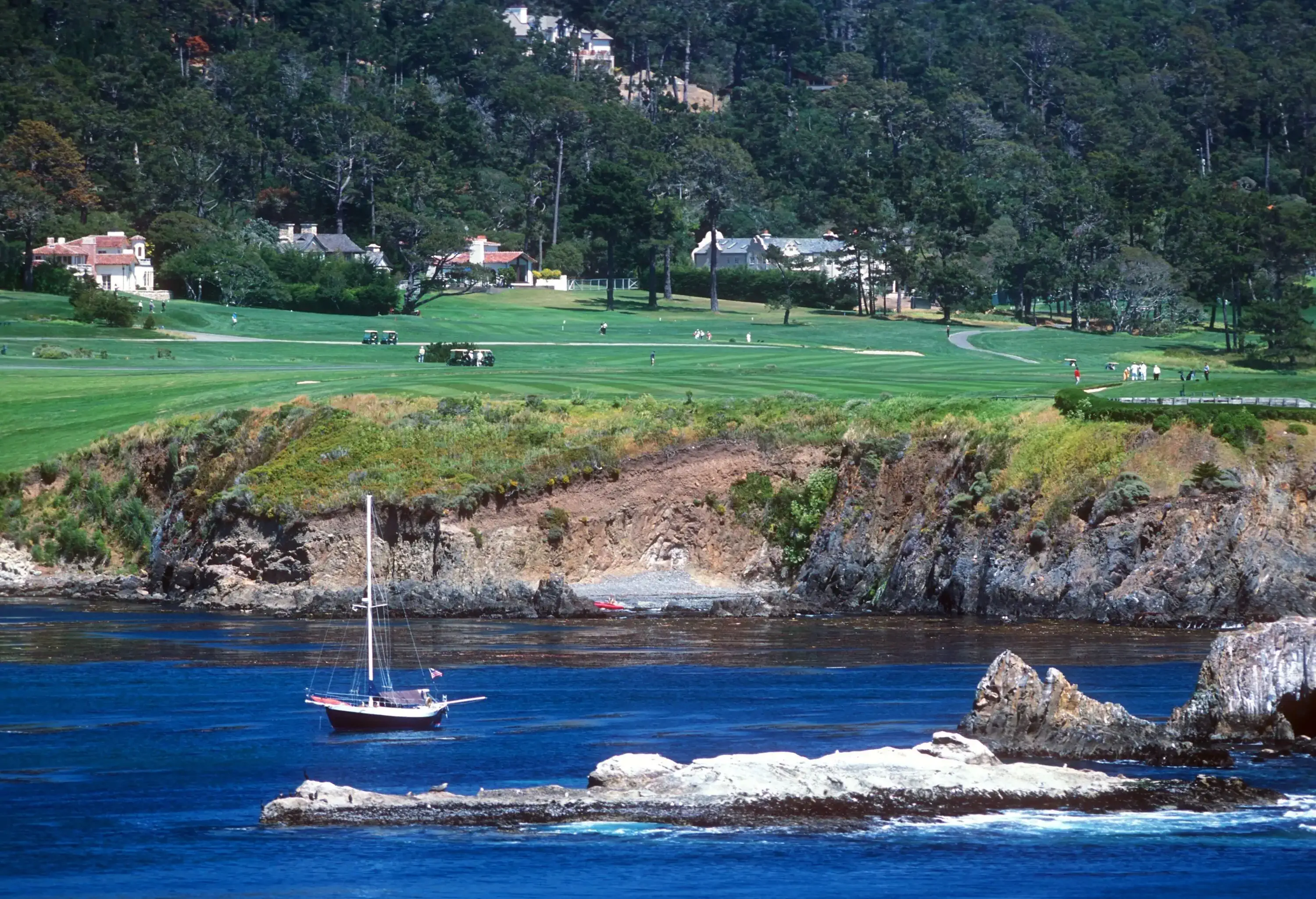 A sailboat floating in a bay's azure waters, with a golf resort in the backdrop.