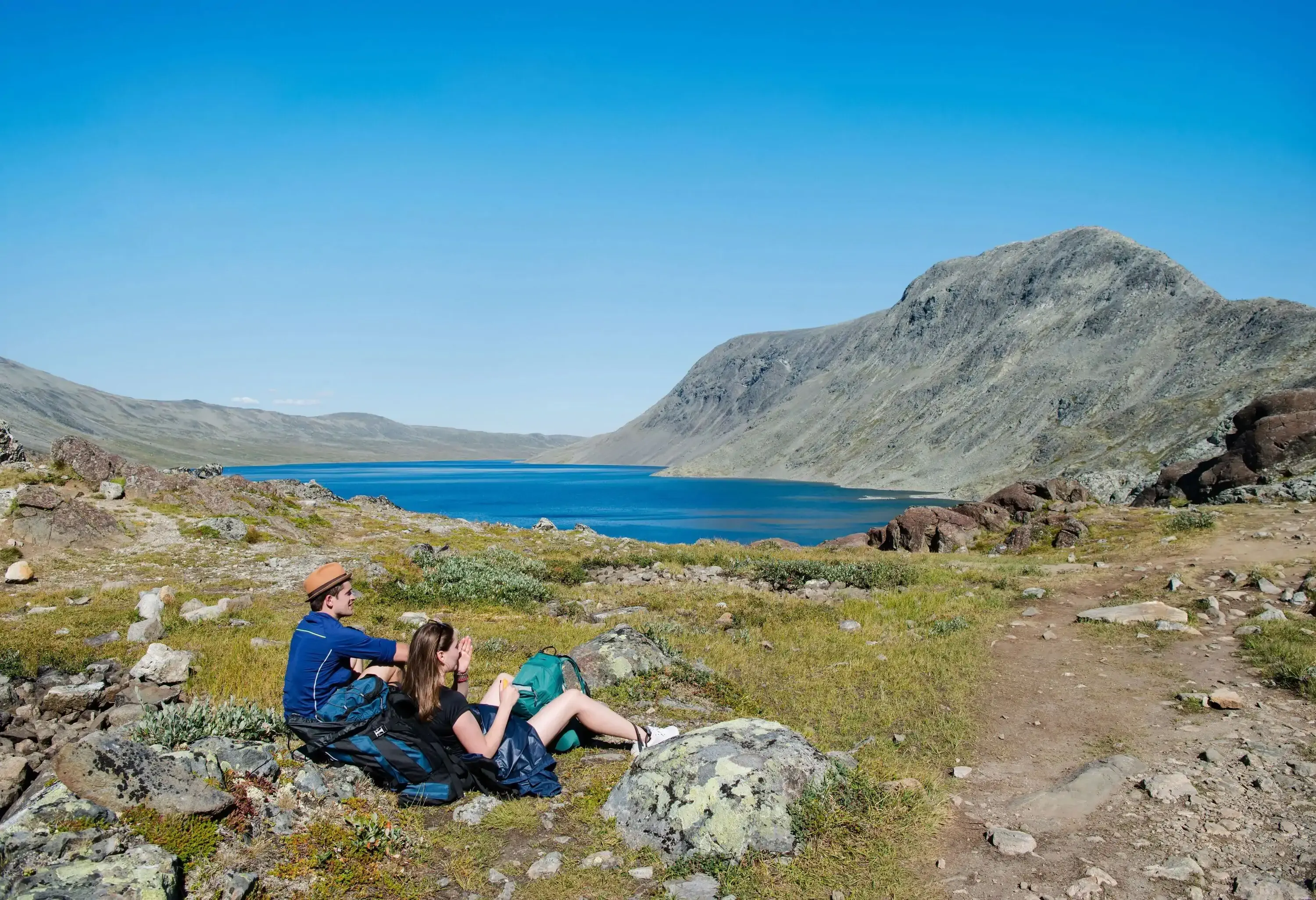 Two young hikers sitting on the shore of a lake surrounded by rocky mountain ranges.