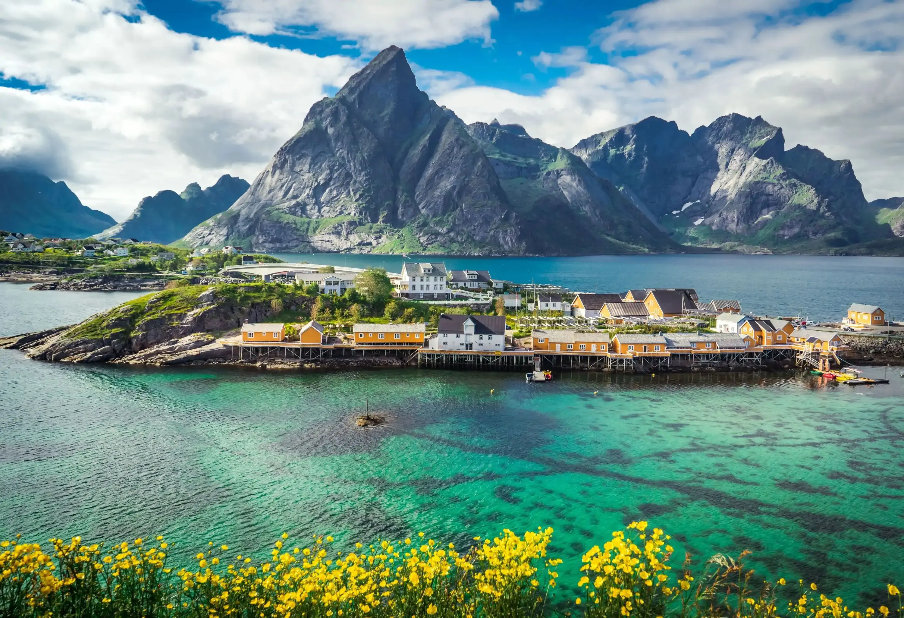 A cluster of yellow traditional houses on stilts in a coastal village against the backdrop of rugged mountains as seen from a hillside with yellow flowers.