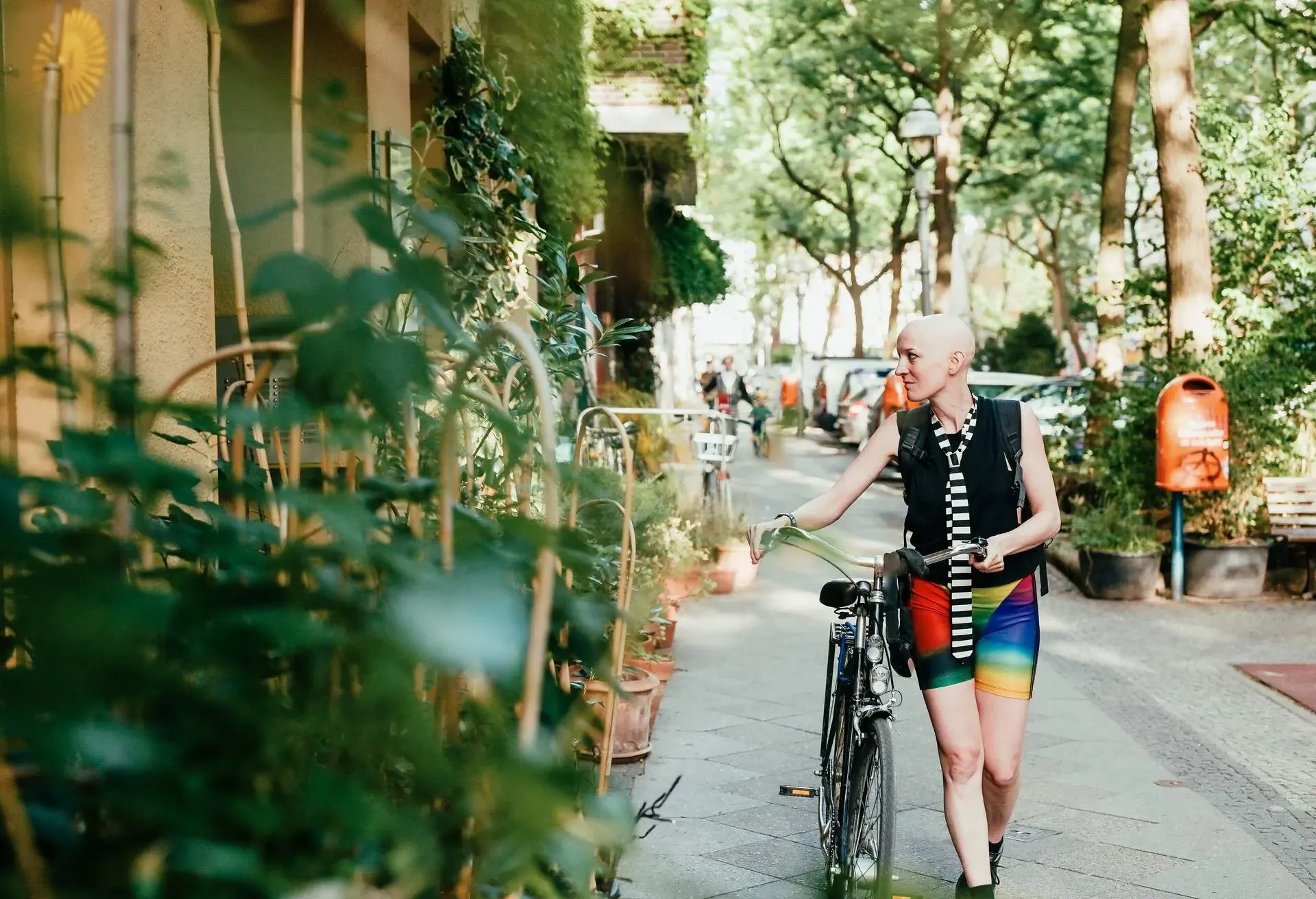 A bald individual with a bicycle in a street lined with buildings adorned with green plants and tall green trees.