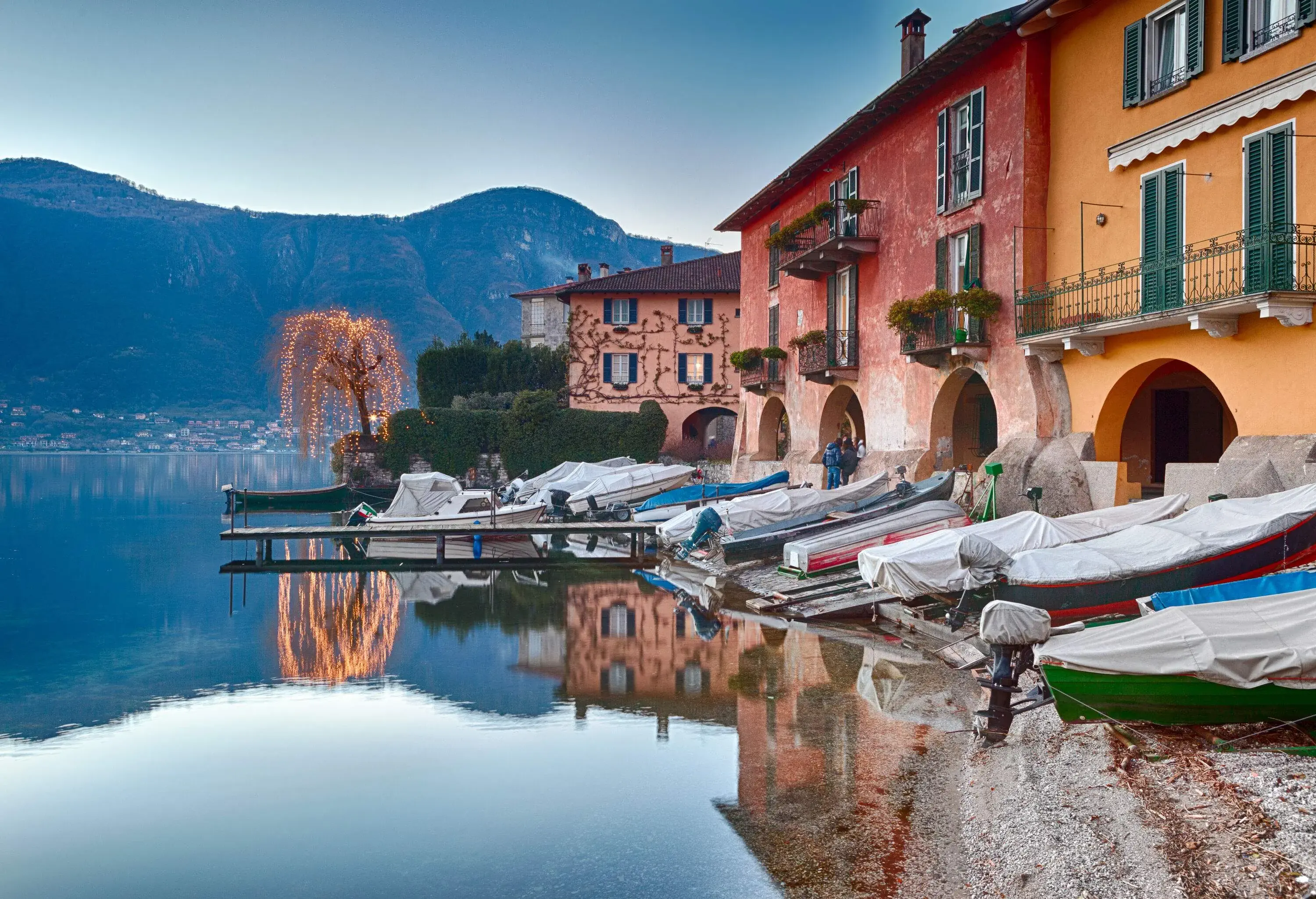 A row of covered boats docked alongside the coastal buildings on a lakeshore.
