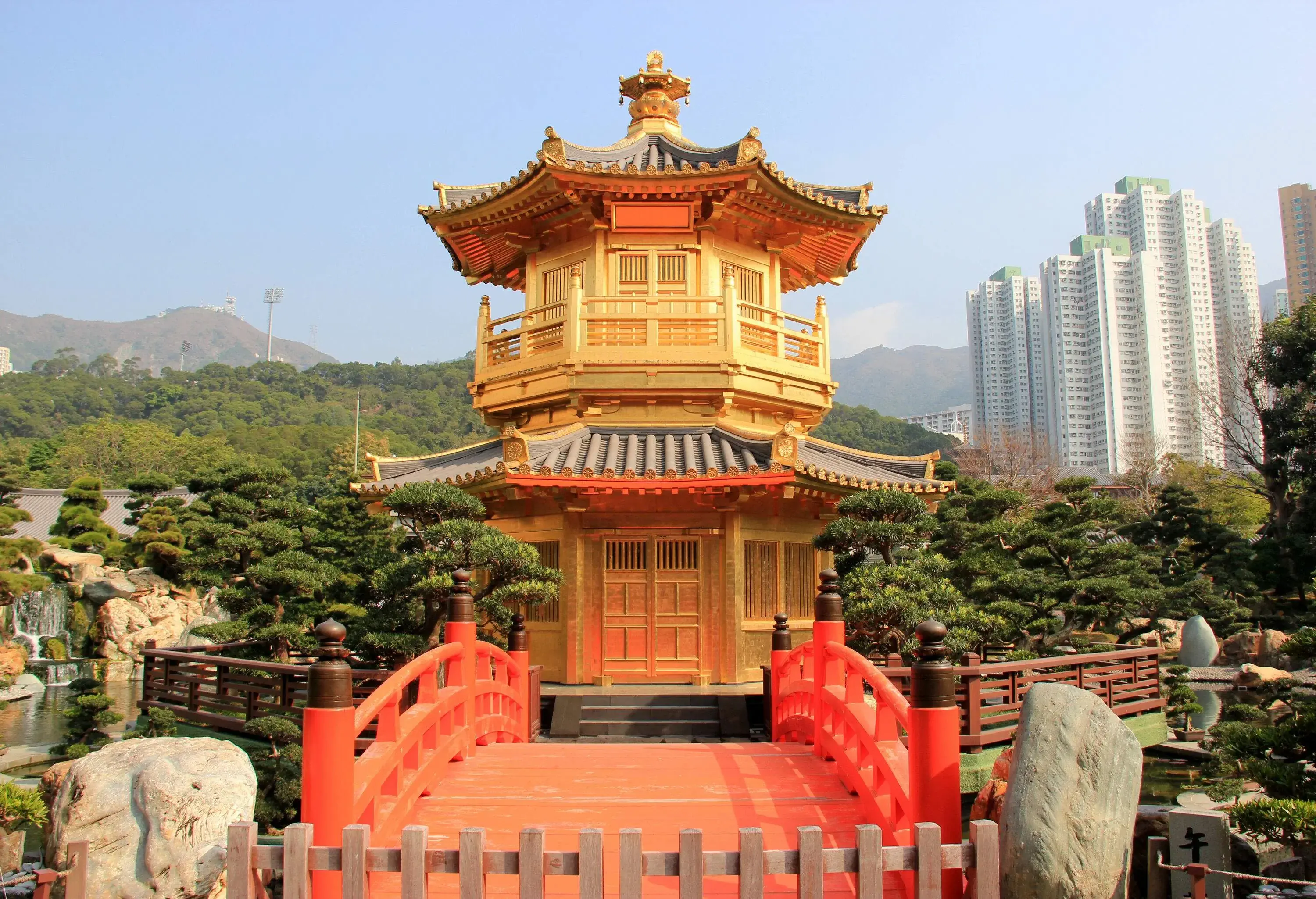 A two-storey golden pavilion accessed through a red bridge in a lush classical Chinese garden.