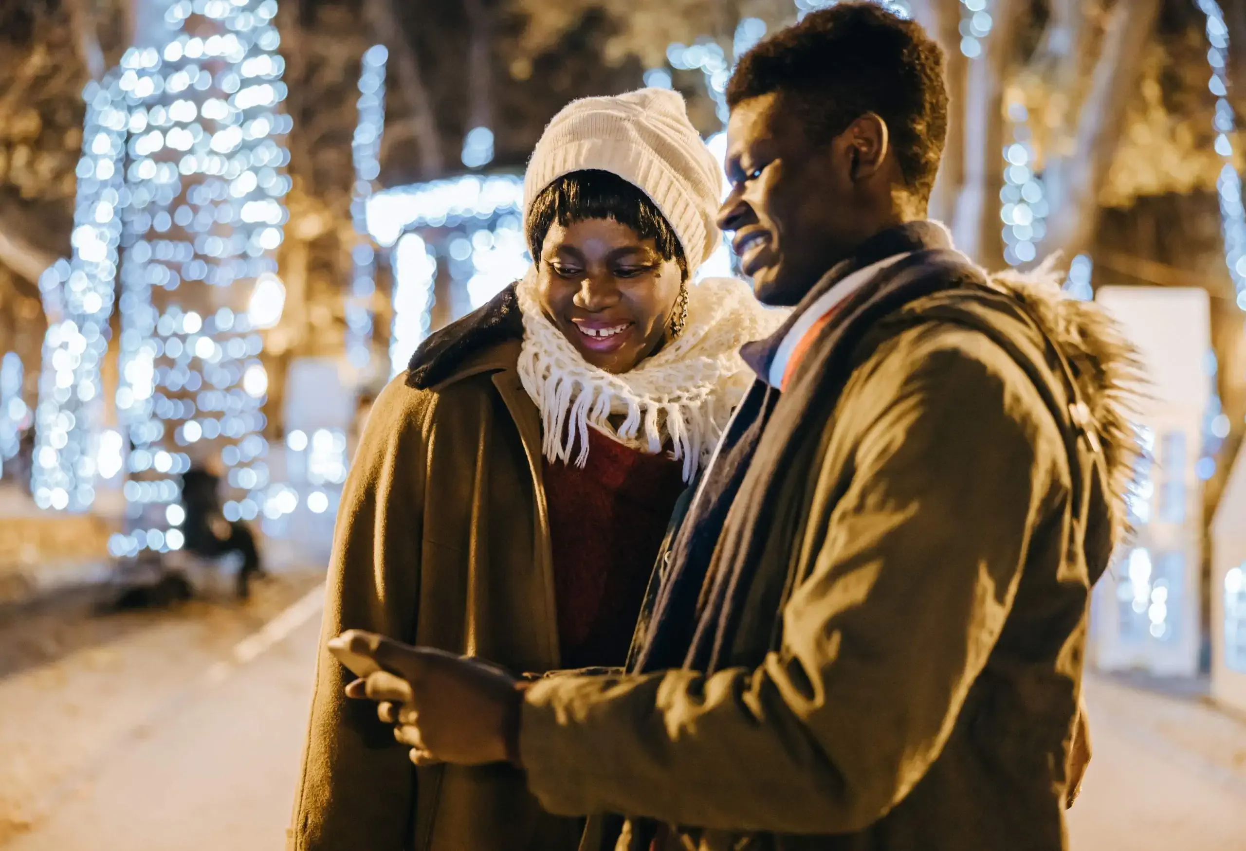 Trees strung with lights serve as a backdrop for a happy young African-American man showing his phone to his mother.