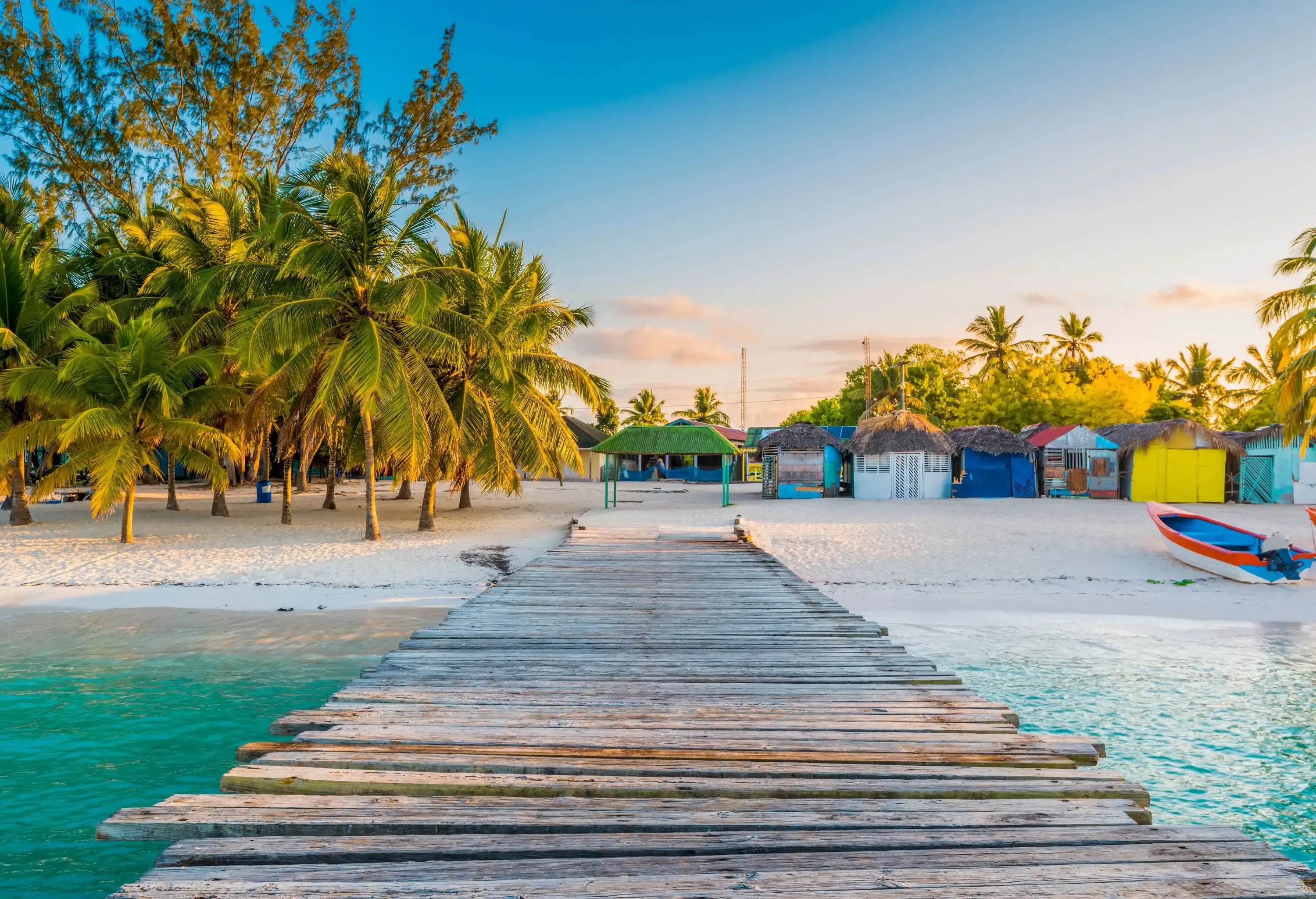 A boardwalk stretching over the turquoise sea to a sandy white beach dotted with coconut trees and colourful cabins.