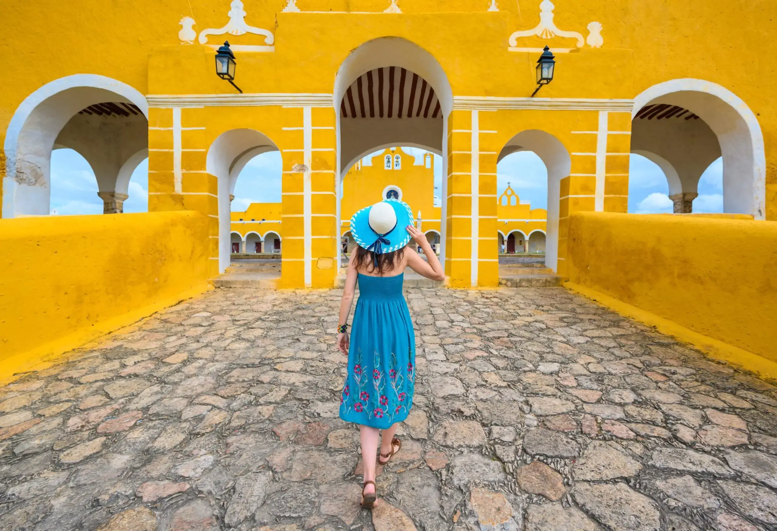 A woman in a blue dress and wide-brimmed hat walks towards a yellow building with multiple arch entrances.