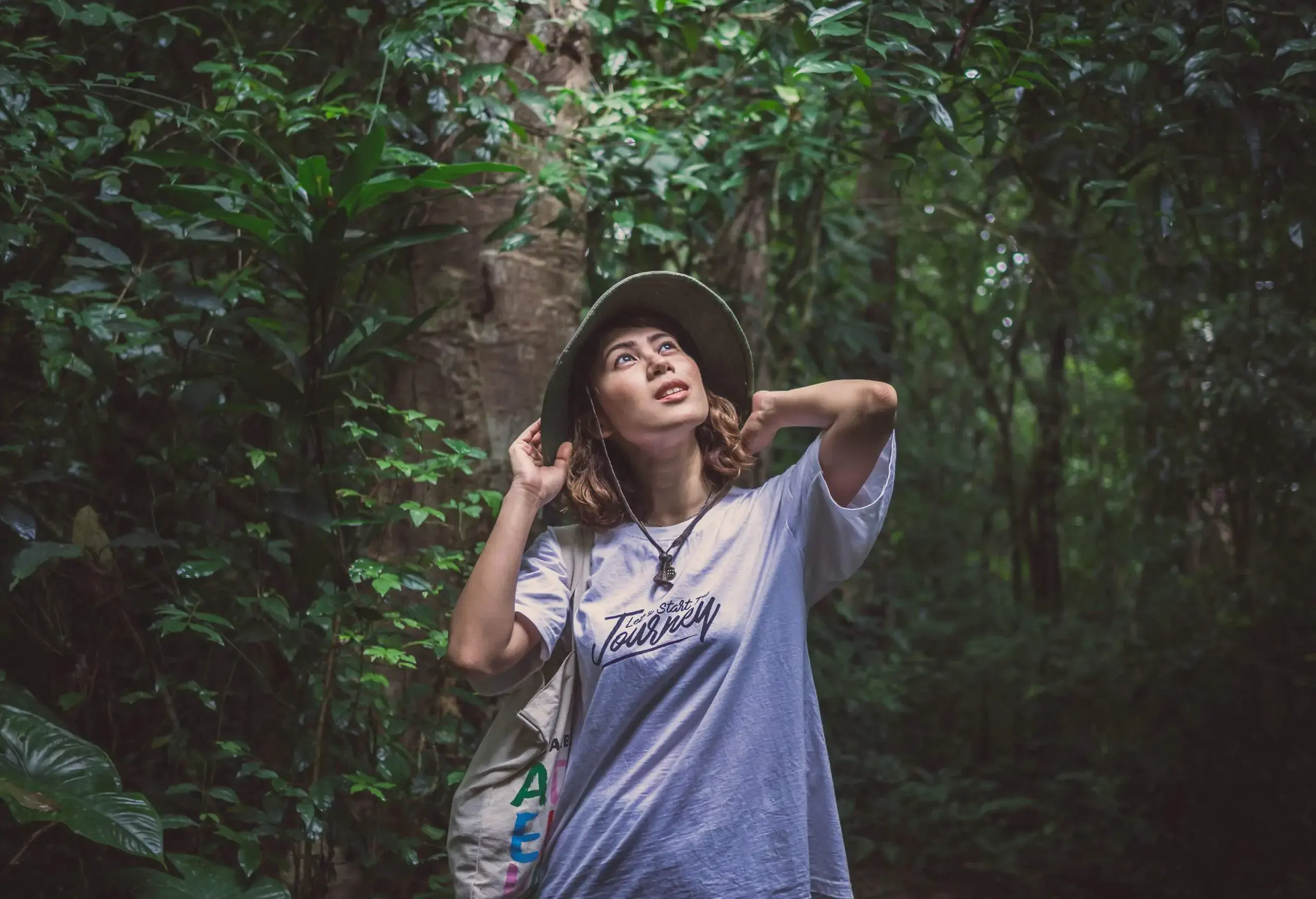 A woman wearing a white shirt, a green bucket hat, and a canvas tote bag looking at a green forest.