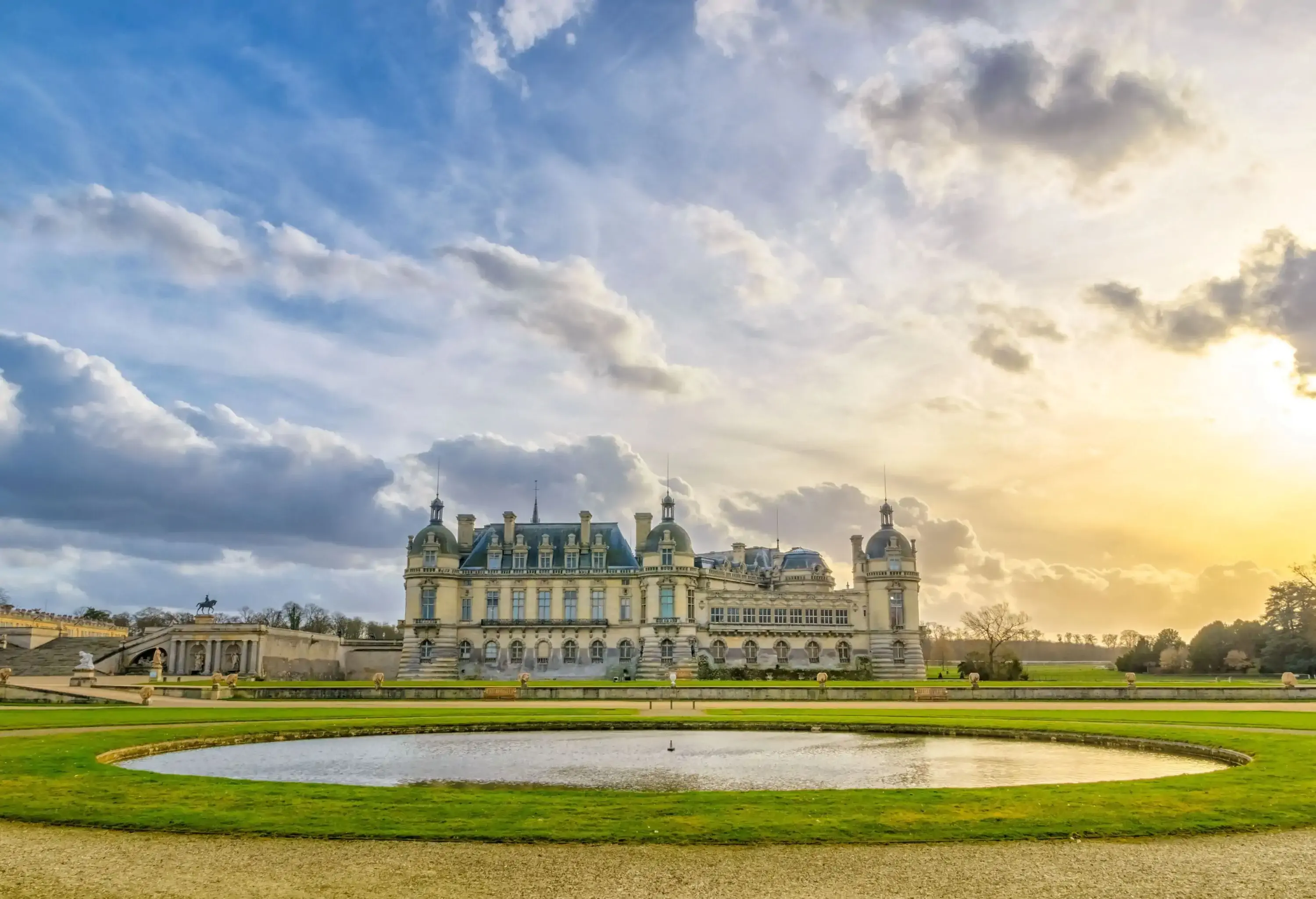 Chantilly Castle, a French Renaissance chateau with blue roofs and beige exterior, behind a water feature on a lawn at sunset.