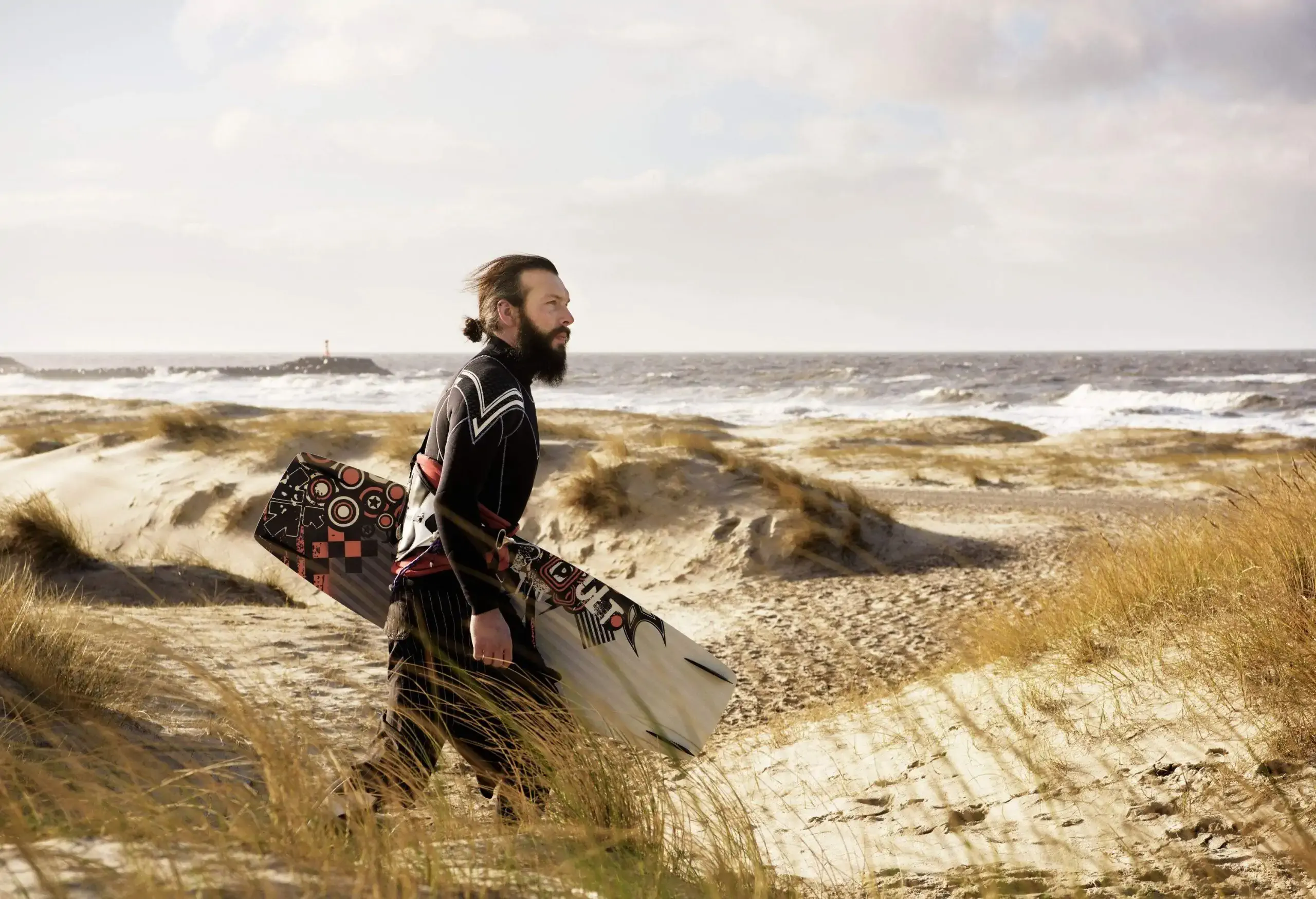 A man carrying a wakeboard walking across the beach with tall grass.