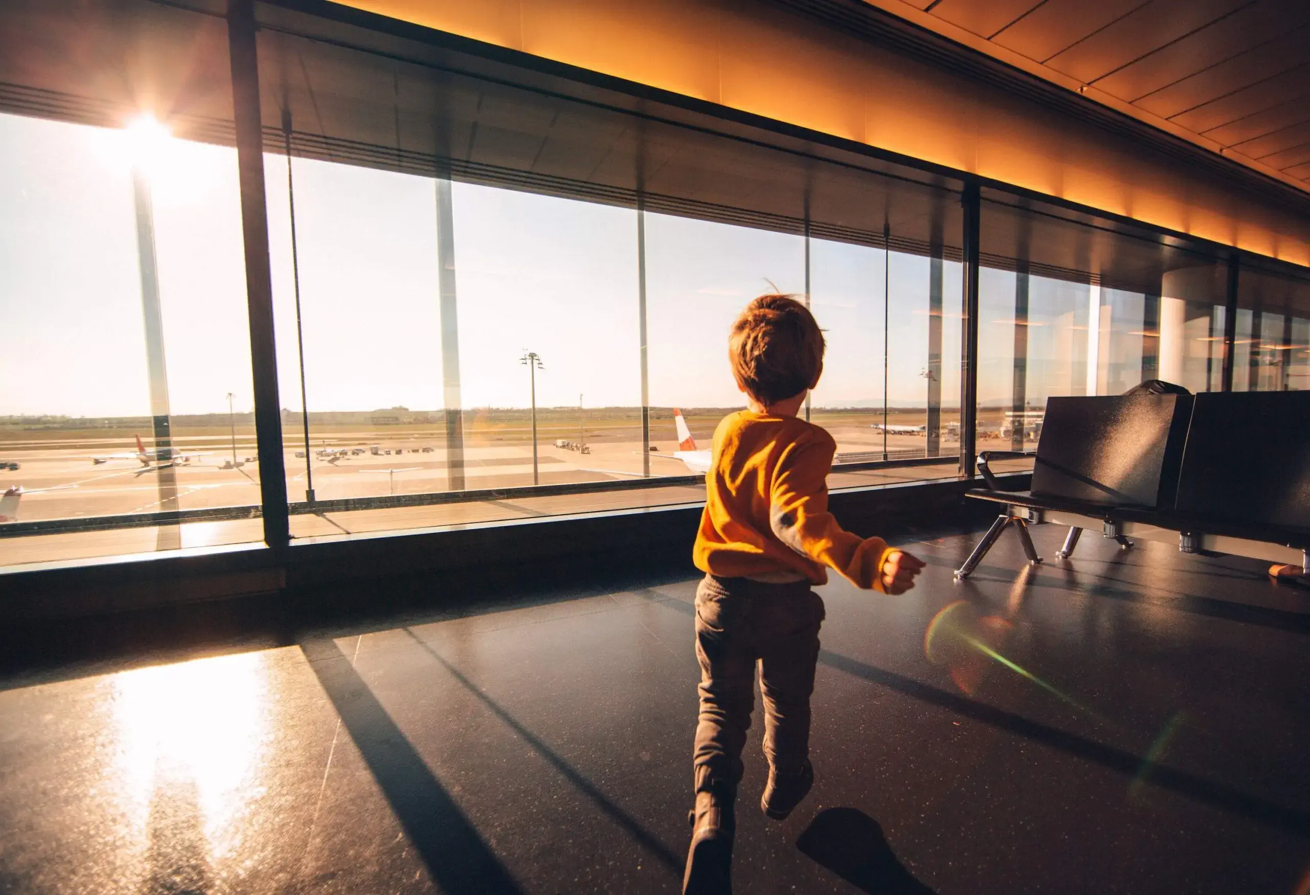 A young child in orange sweater runs on an airport lounge with large windows.