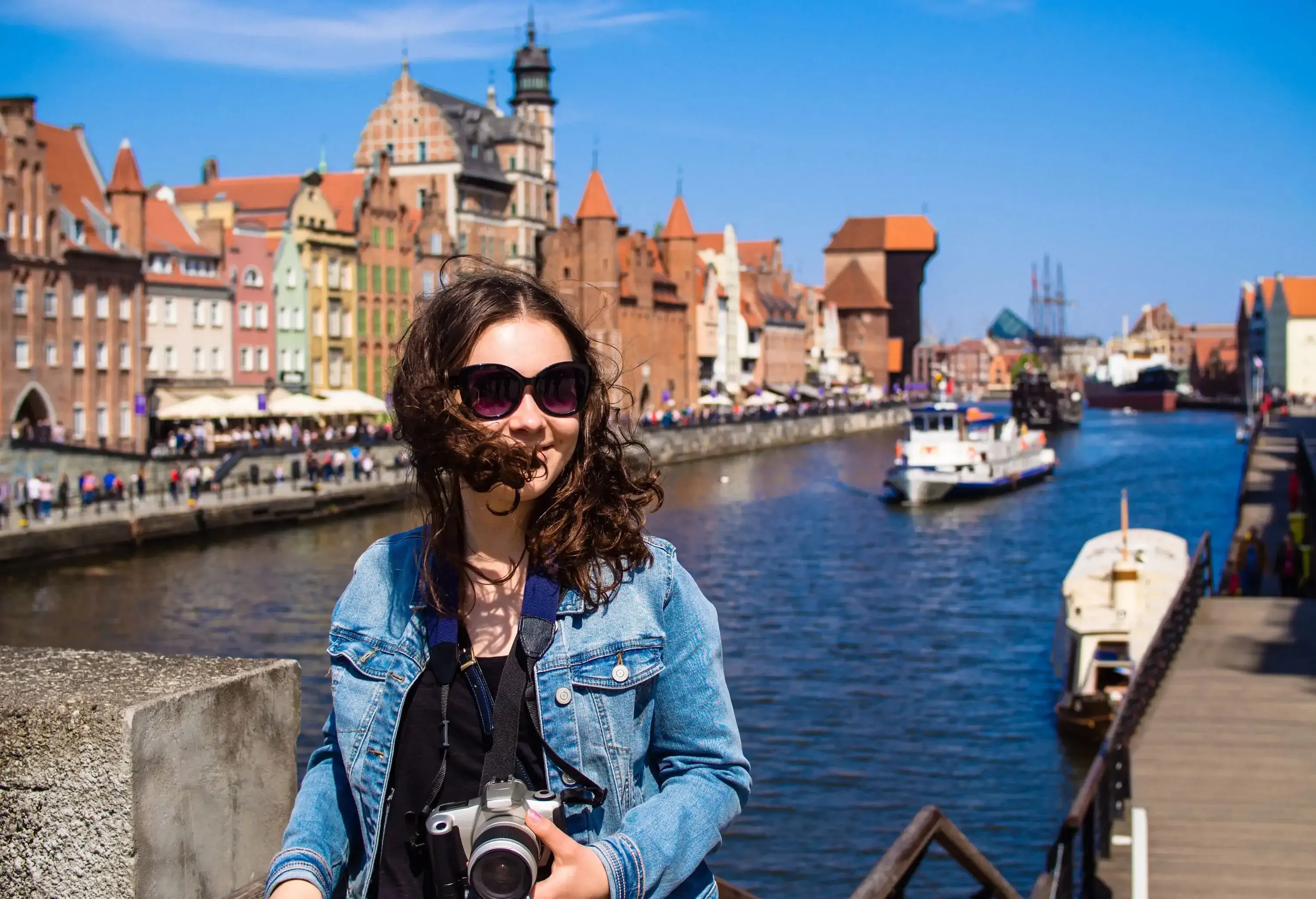 A woman in sunglasses and denim jacket holds a camera as she stands against the backdrop of a water canal along adjacent city buildings.