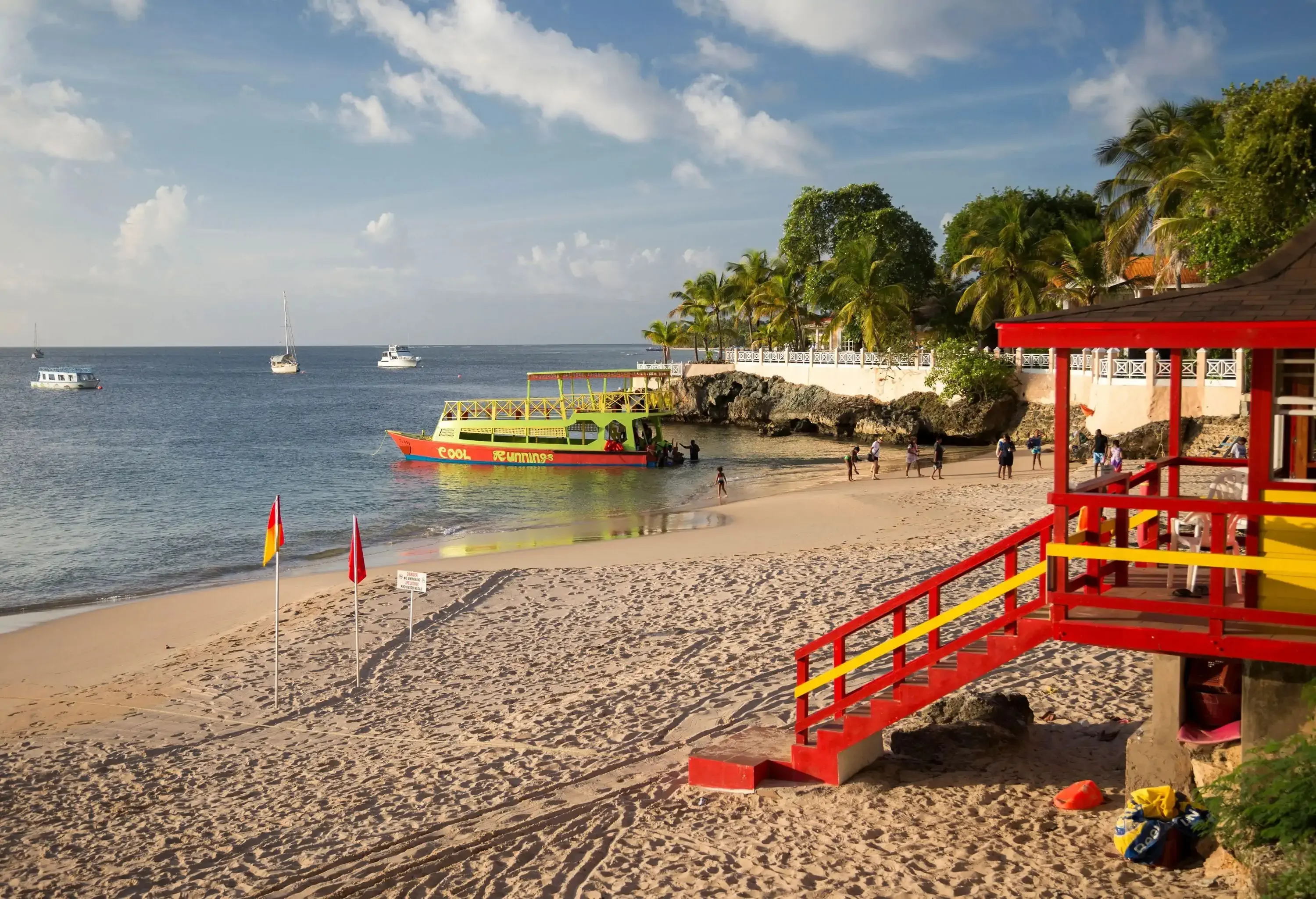 A red lifeguard tower overlooks a beach with tourists disembarking from a boat.
