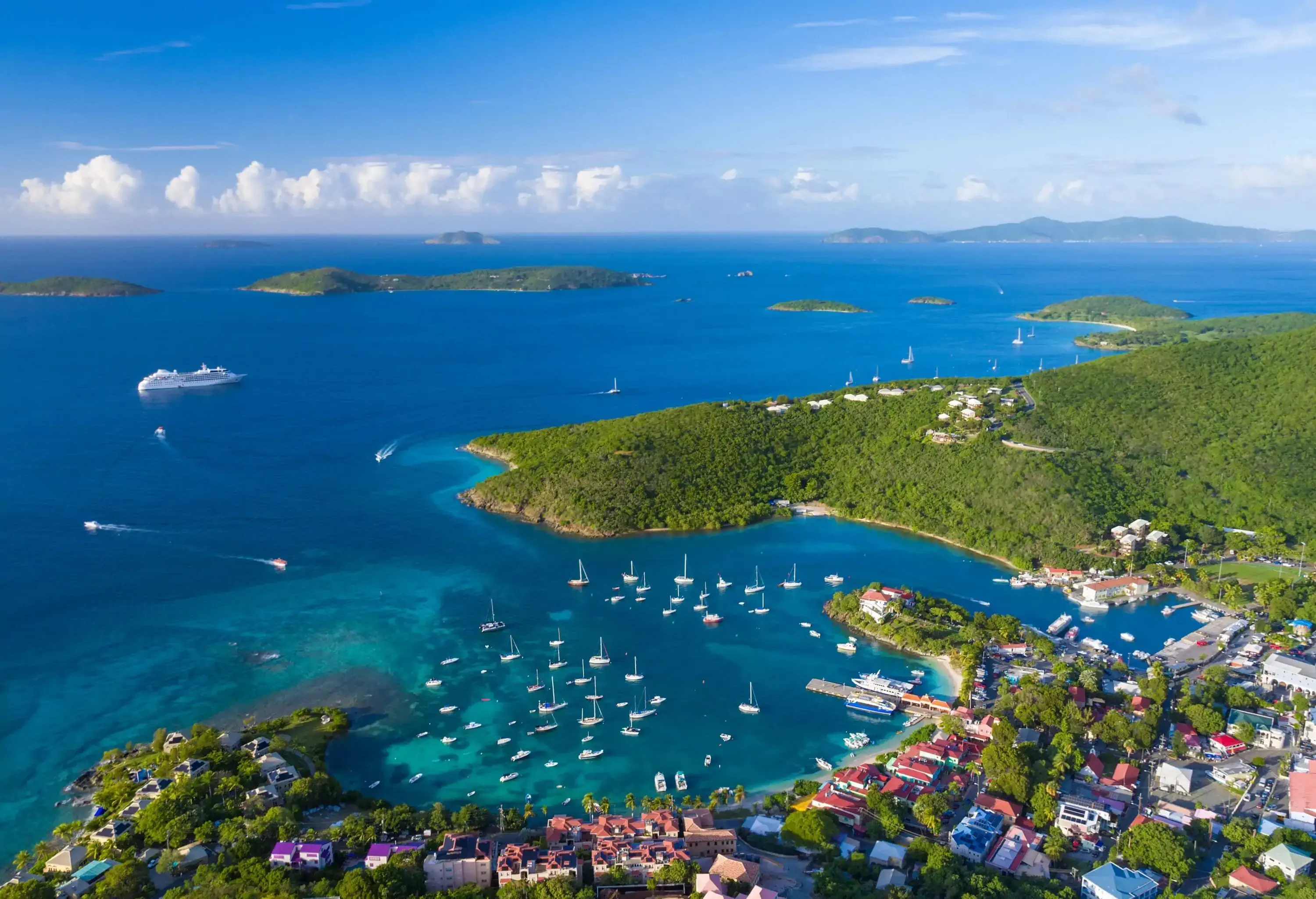 A sheltered cove with anchored yachts along a coastal village with views of small islands in the ocean.