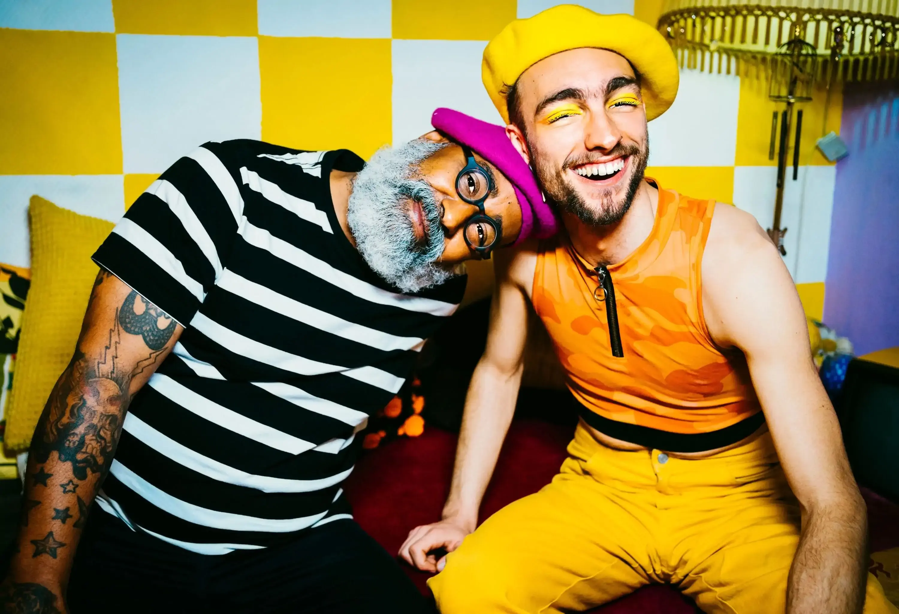 Two men wearing flat-crowned caps sitting together in a brightly coloured room.
