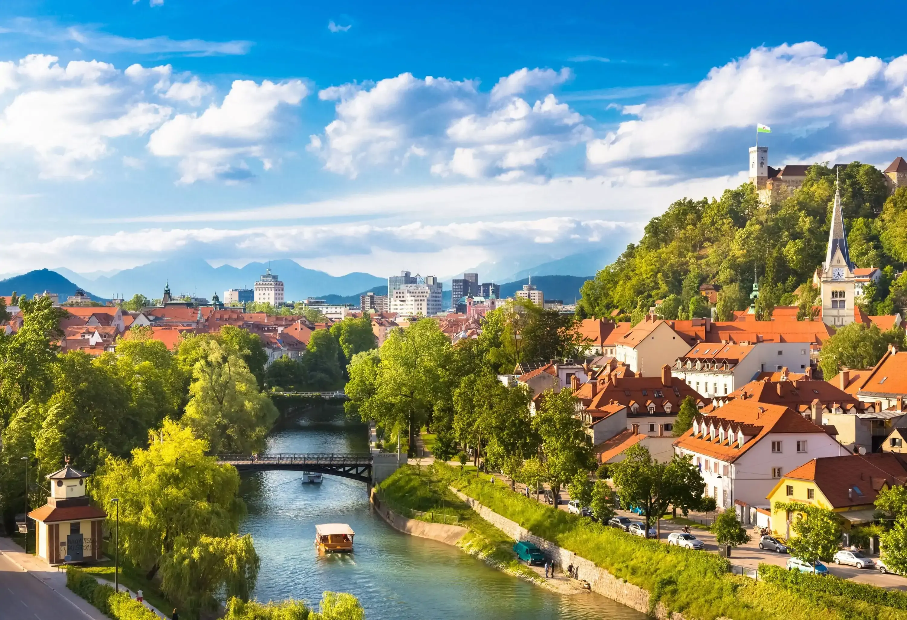 A boat cruising down a river with historic structures and beautiful trees covering the banks and hilltops.