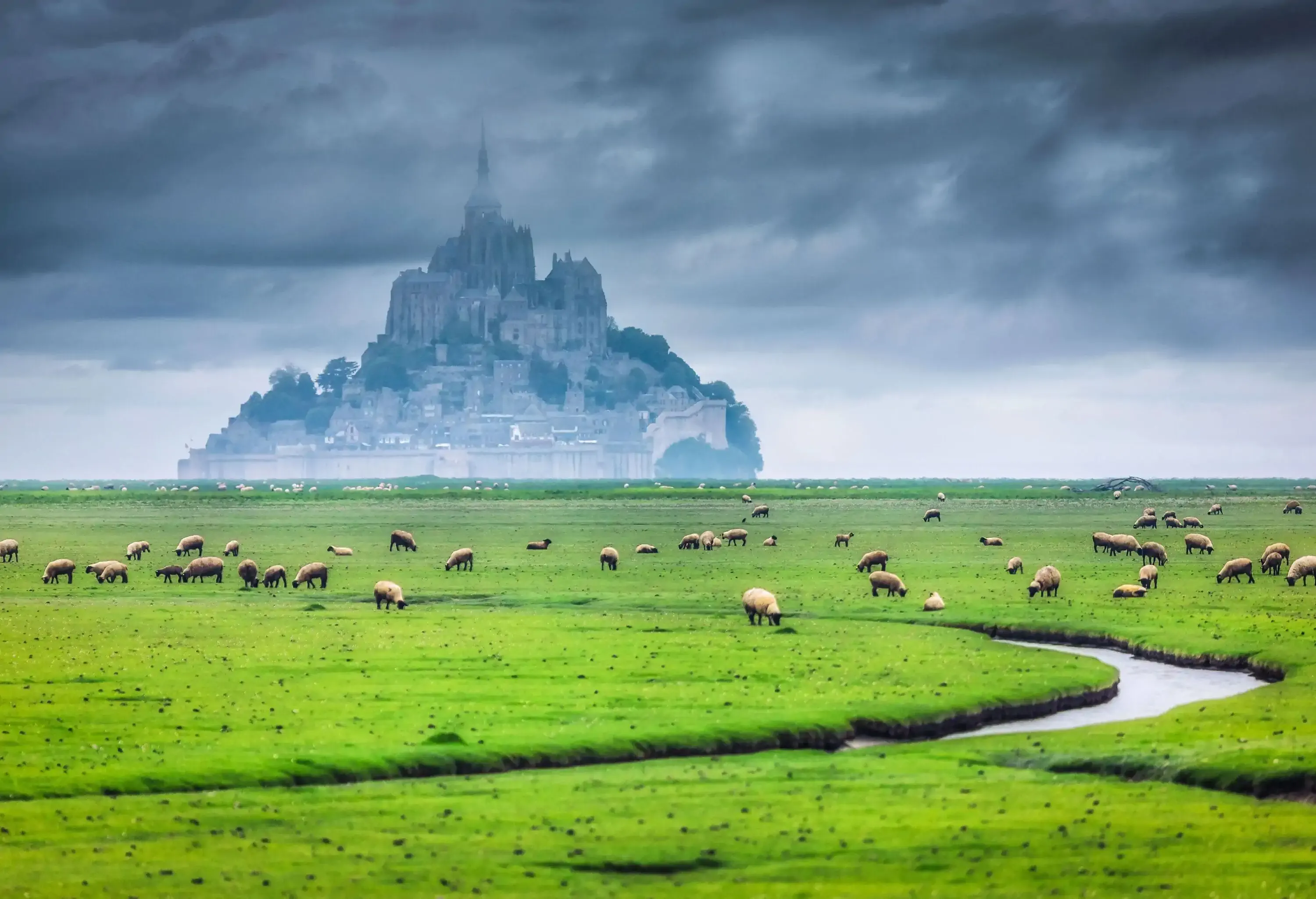 A green pasture with a small stream and grazing sheep and views of a town set on a hill with a steeple on top.