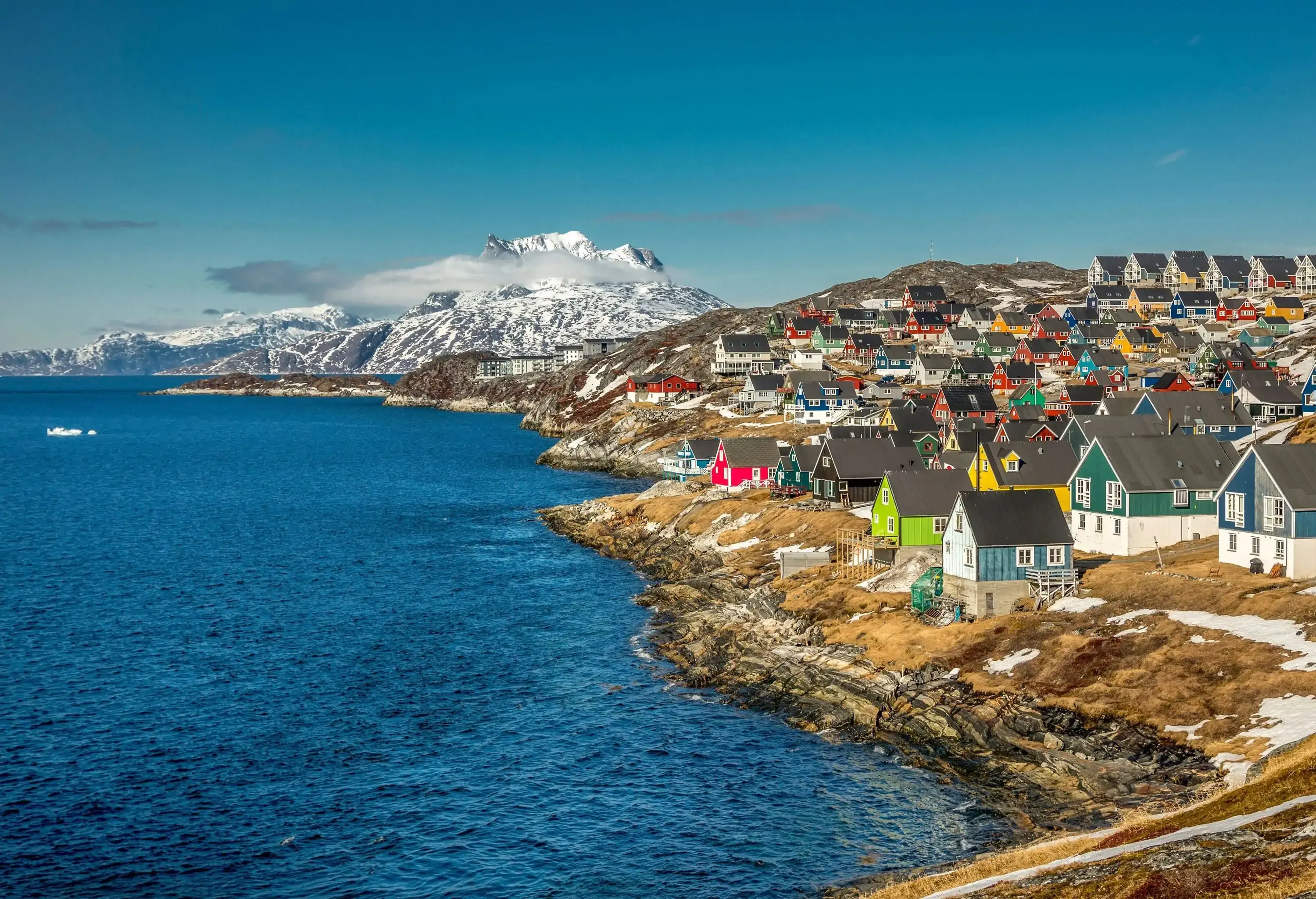 A seaside town with brightly coloured houses on the rocky island against the backdrop of a snow-dusted mountain.
