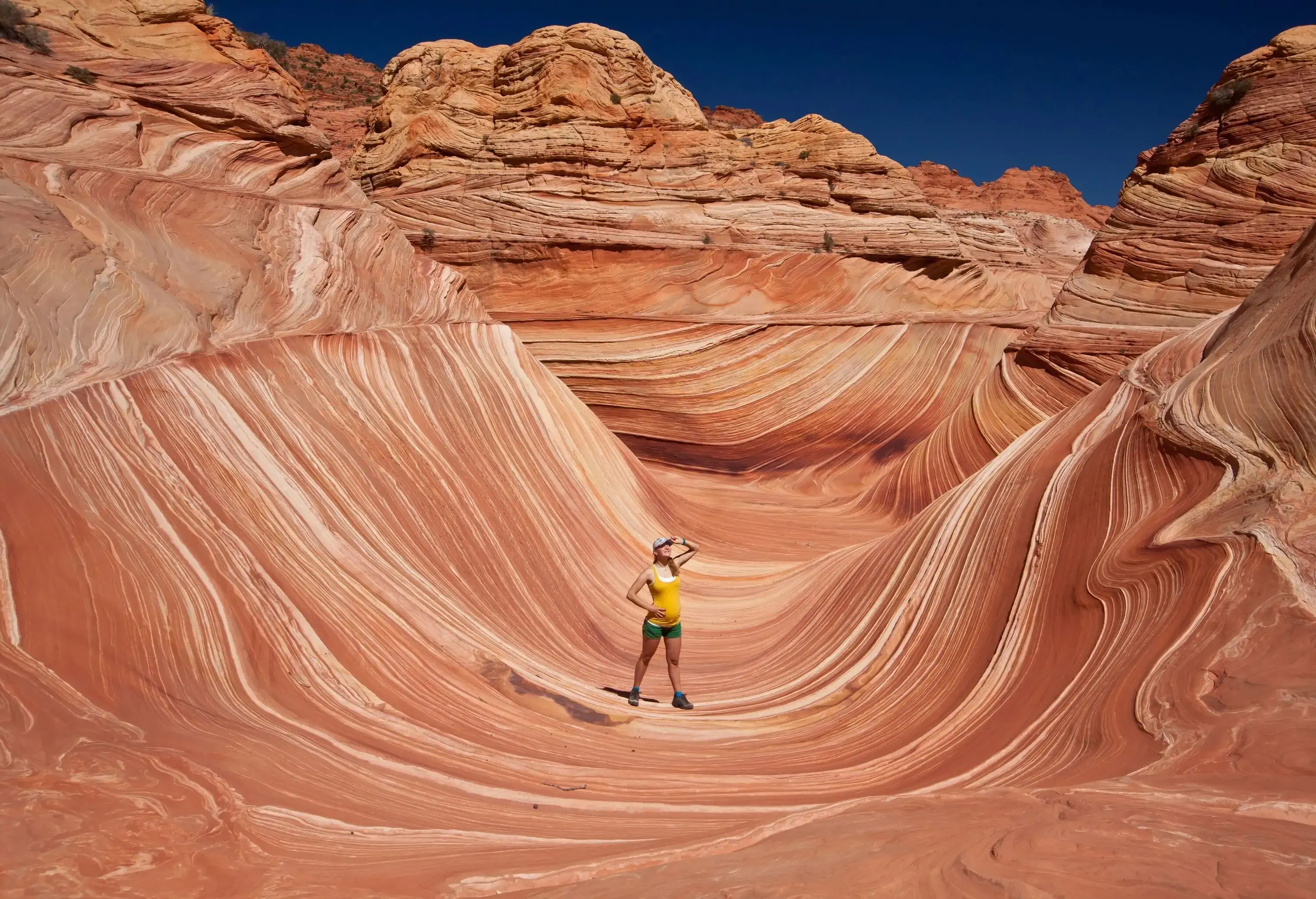 A female hiker in a yellow top and green shorts standing in the middle of The Wave canyon.