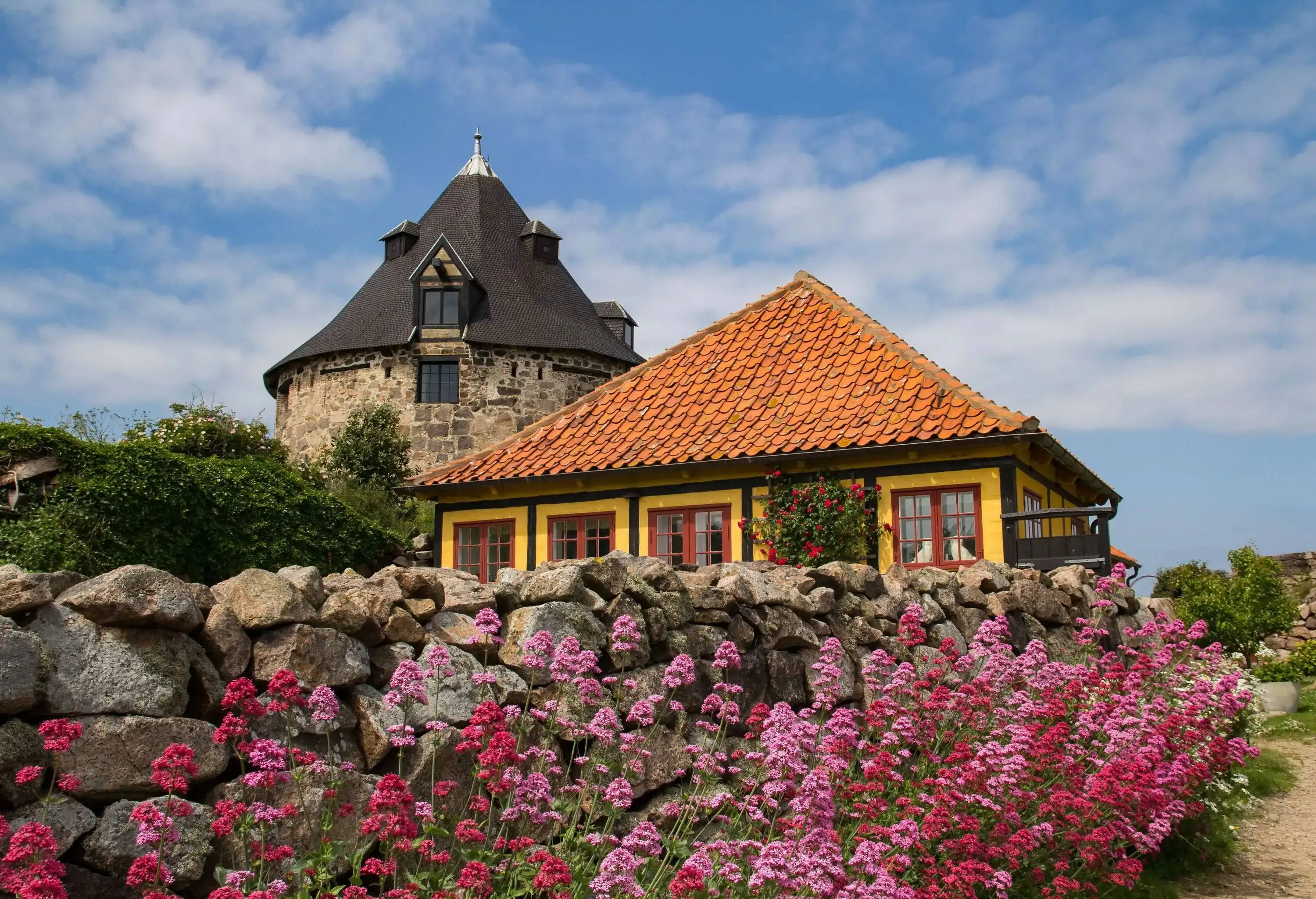 a traditional yellow half-timbered house with a red-tiled roof and a historic round stone tower behind it. The foreground is filled with blooming pink and red flowers.